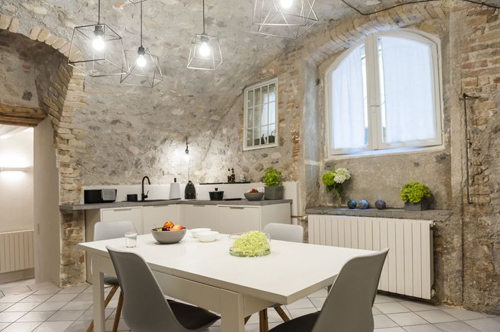Cucina e sala da pranzo con pareti in mattoni a vista, tavolo bianco e sedie grigie. Finestre luminose e lampade a sospensione.