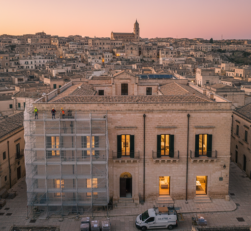 Facciata di un edificio con impalcature, operai, finestre illuminate. Sullo sfondo il paesaggio urbano al tramonto.