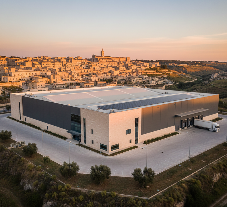 Ampio magazzino moderno con pannelli solari, con vista sulla città al tramonto. Un camion attracca.