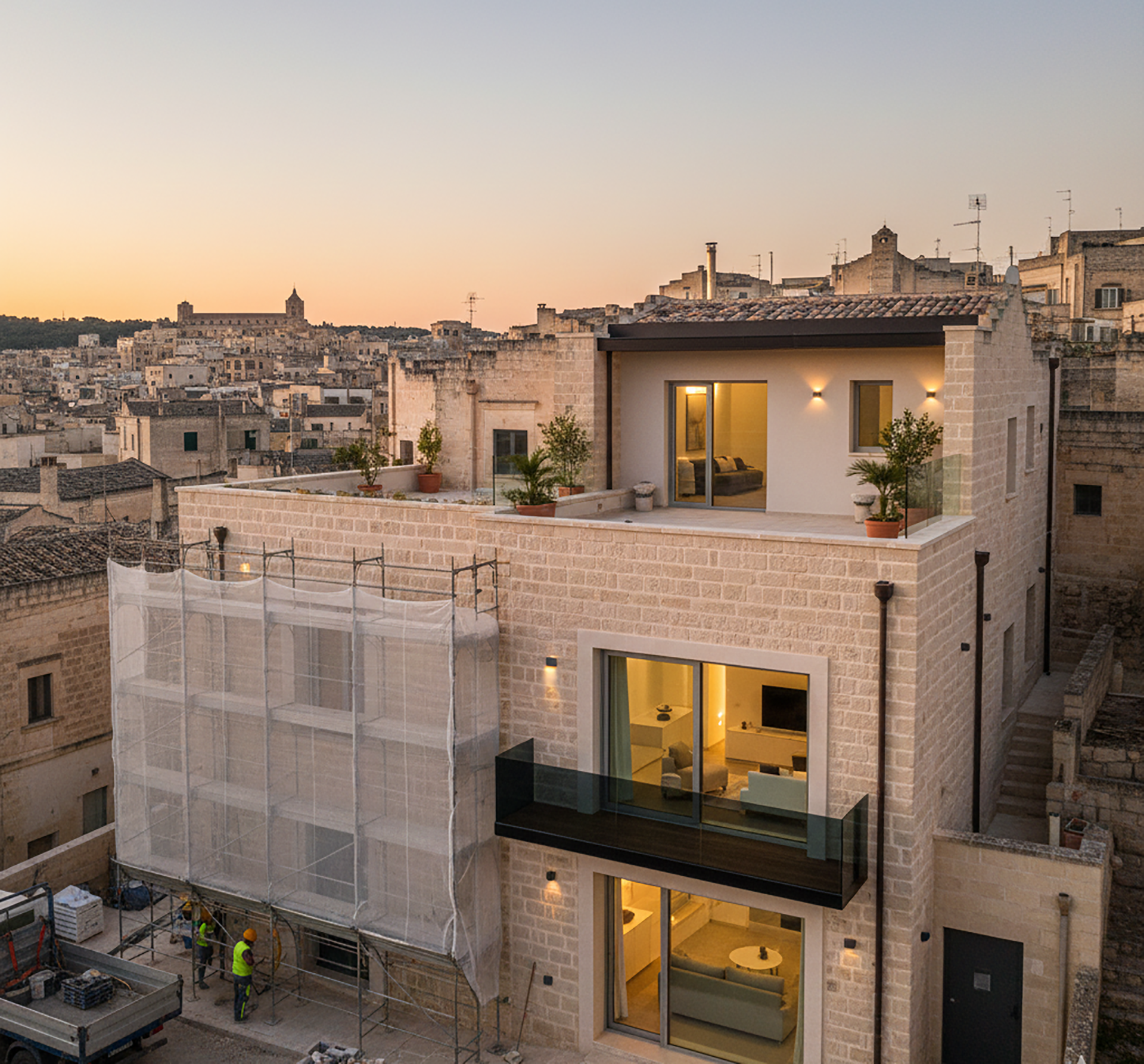 Edificio in pietra con aggiunte moderne, situato in un centro storico. Luce serale, impalcature laterali.