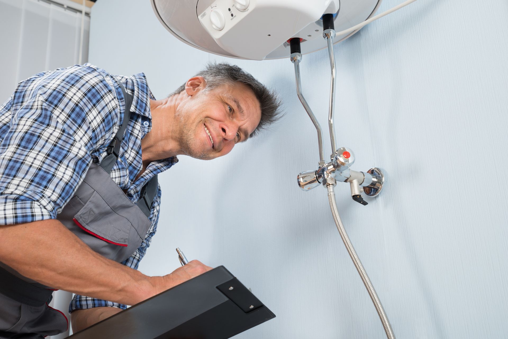 Plumber checking a water heater, writing on a clipboard. Smiling in a light-blue room.