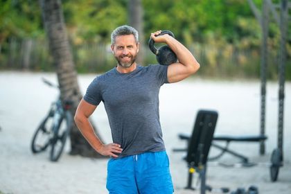 Man with grey hair, holding a kettlebell overhead, smiling on a beach.