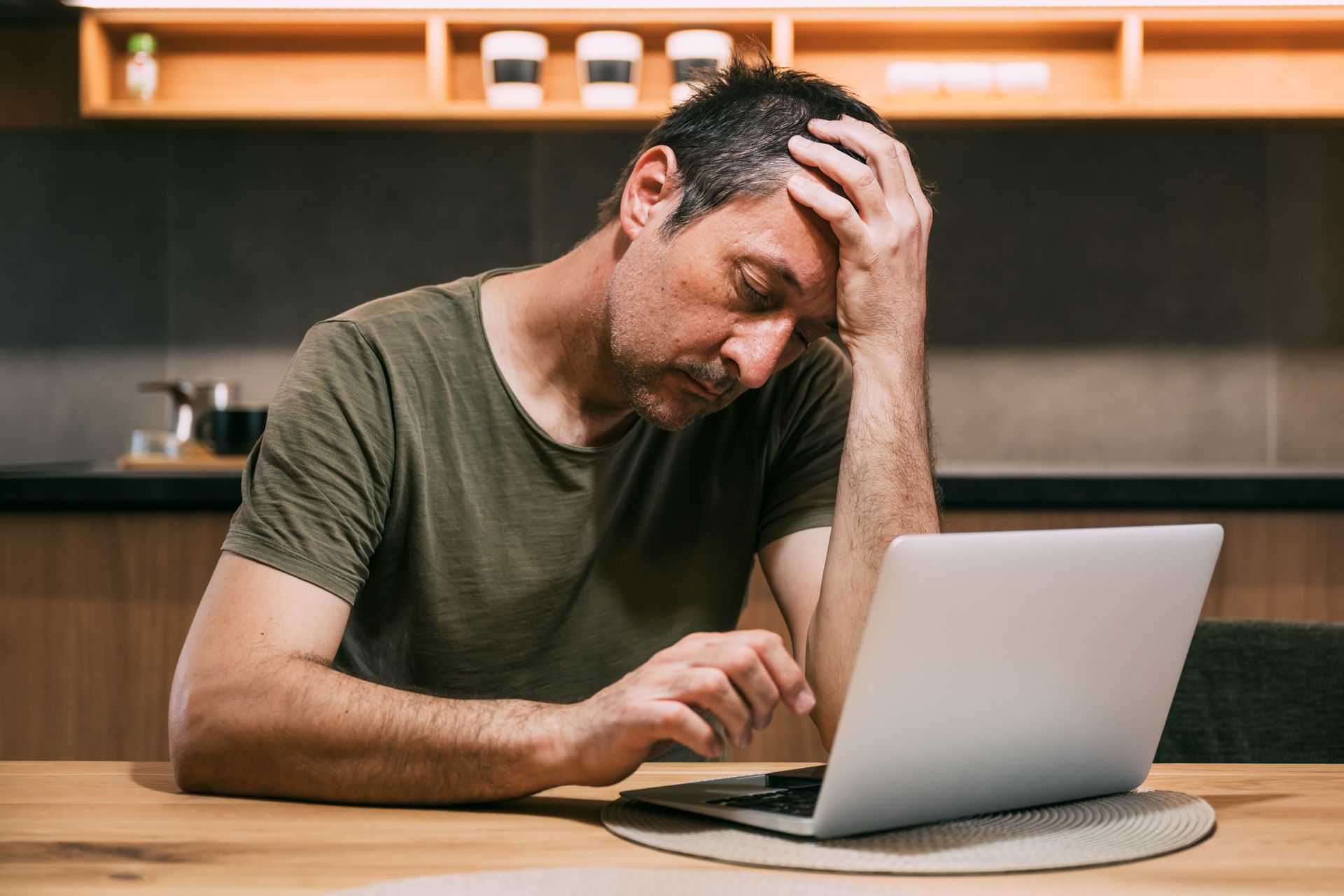 Man with hand on forehead looks frustrated, sitting at a table with a laptop in a kitchen.