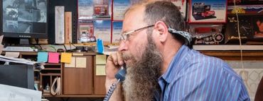 A man with a beard is sitting at a desk talking on a phone. | White Sulphur Garage