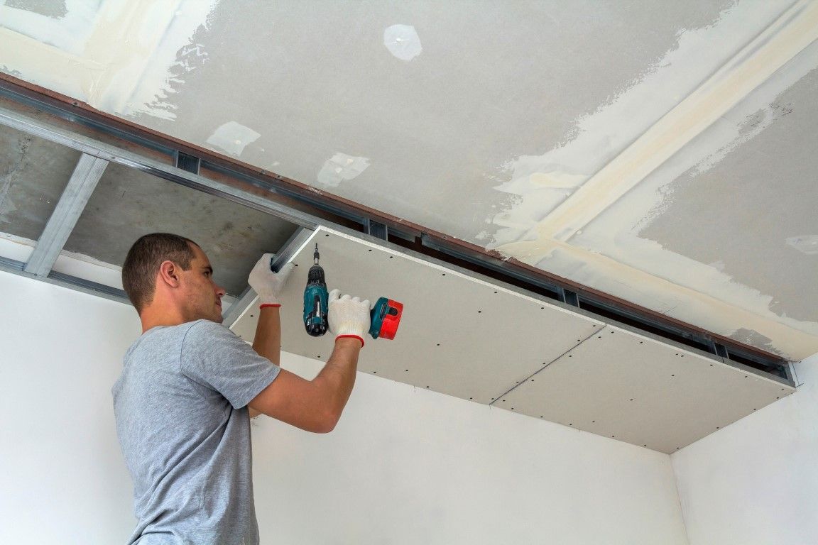 A person wearing protective gear and a respirator uses a paint sprayer on a wall in a room covered in plastic sheeting.