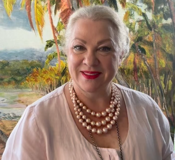 A woman wearing a pearl necklace is smiling in front of a painting.