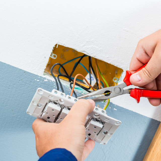 Hands wiring a white electrical switch into a wall with exposed wires and pliers.