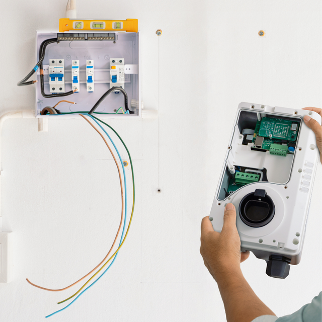 Person installing EV charger, wires exposed near electrical panel on white wall.