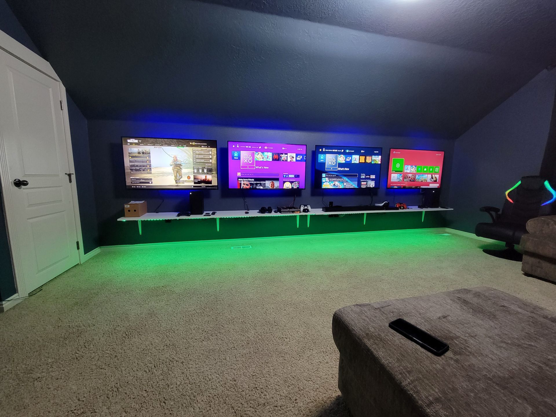 Gaming room with four TVs, white shelves, green LED lights, and a gaming chair.