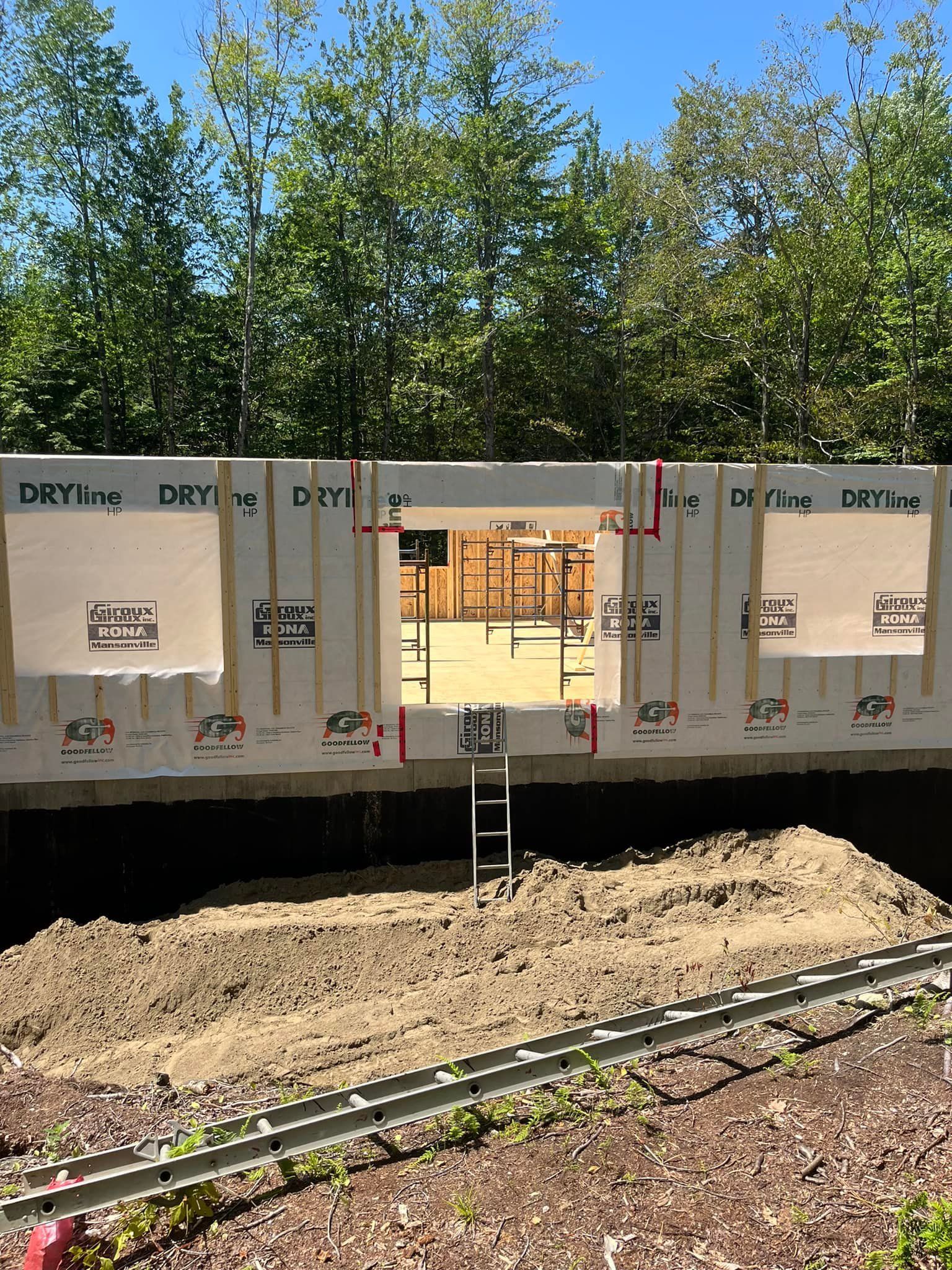 Chantier de construction avec charpente apparente, ouvertures pour fenêtres, échelle dans une tranchée.