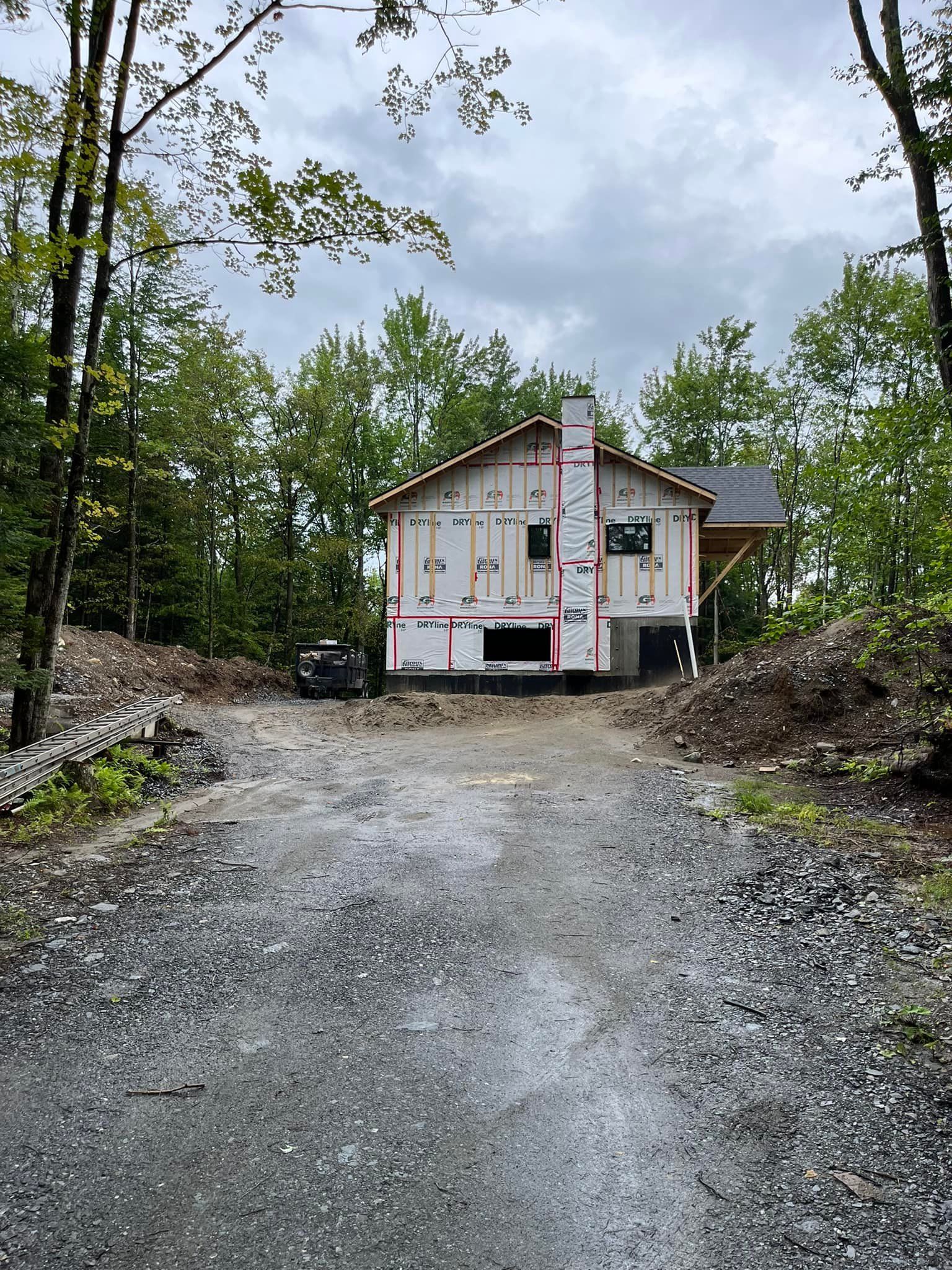 Allée de gravier menant à une maison en construction dans une zone boisée ; ciel couvert.