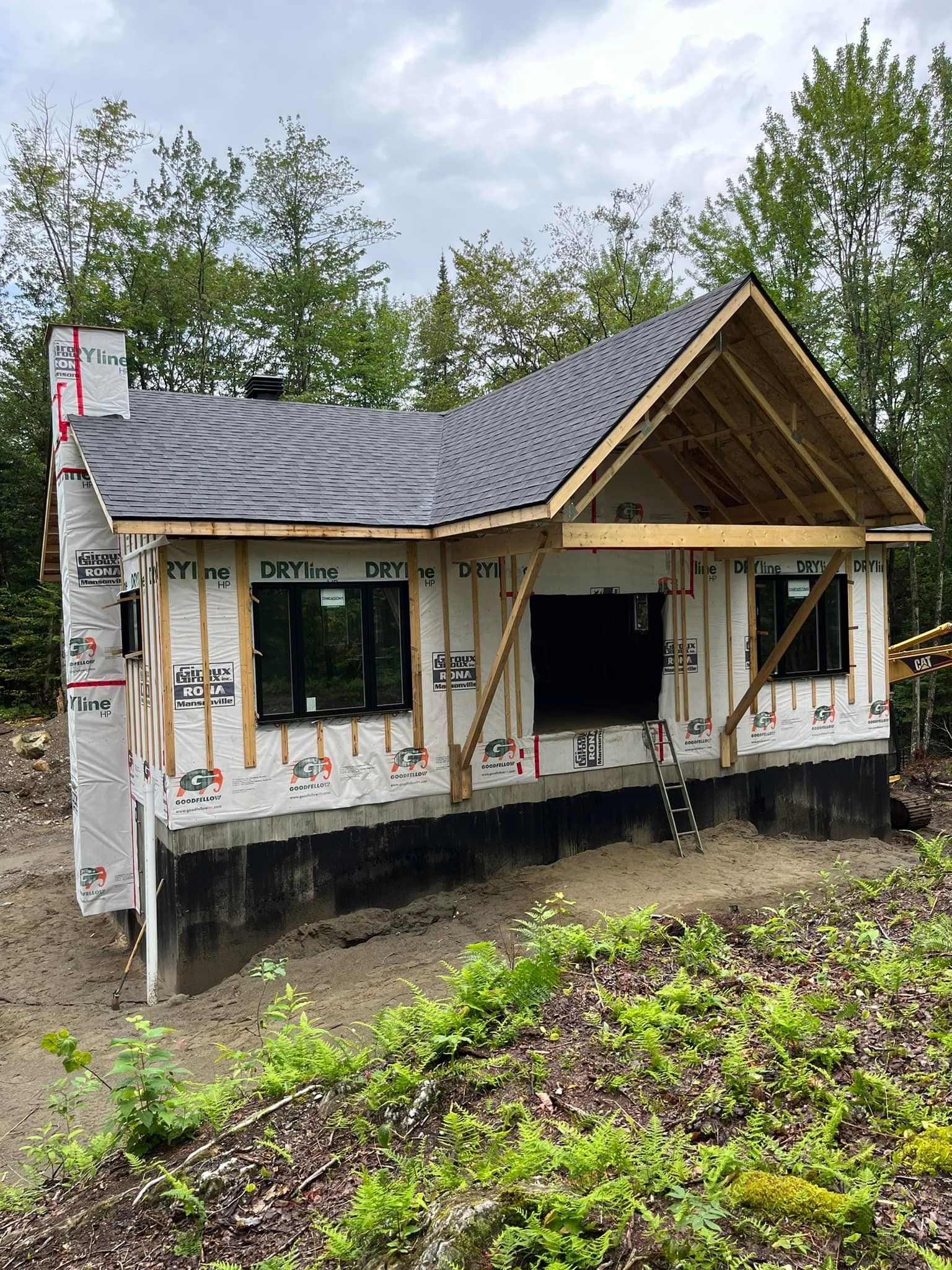 Un bâtiment en construction partielle, avec un toit sombre et une cheminée, entouré d'arbres et de terre.