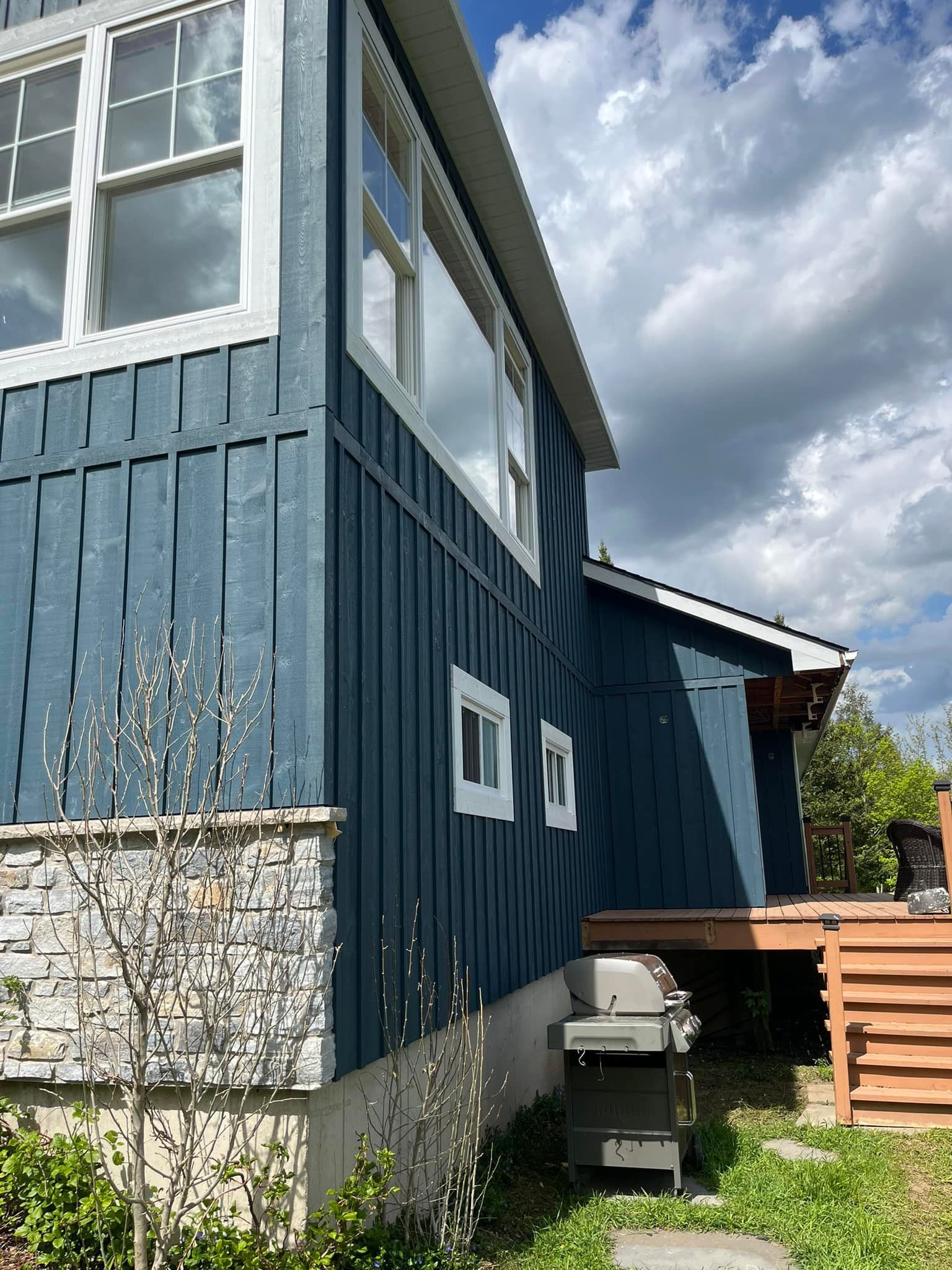 Maison aux murs bleus, fenêtres à encadrement blanc, fondations en pierre et barbecue par une journée ensoleillée.