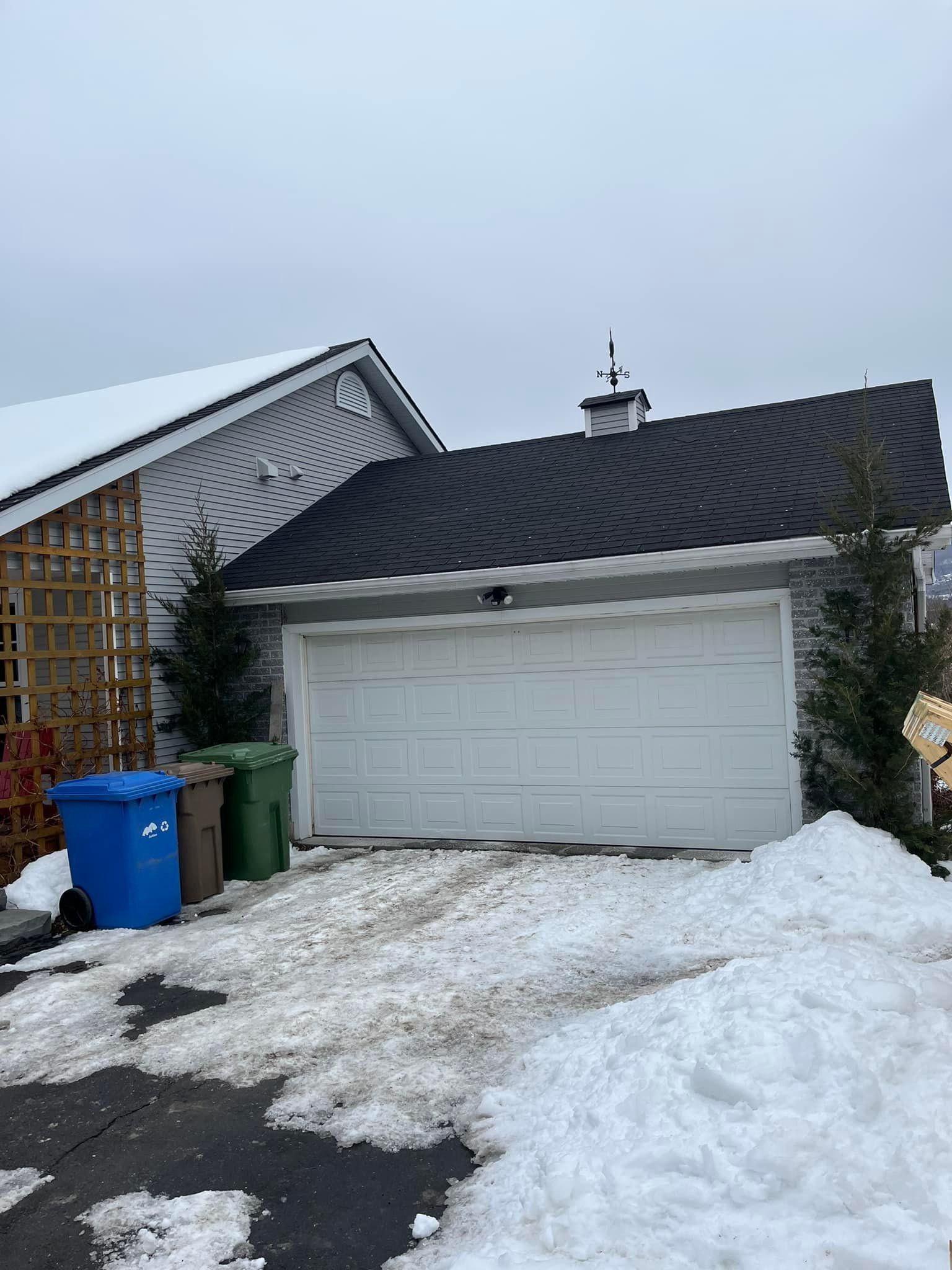 Garage avec porte blanche, allée enneigée, poubelles bleues, brunes et vertes, et ciel nuageux.