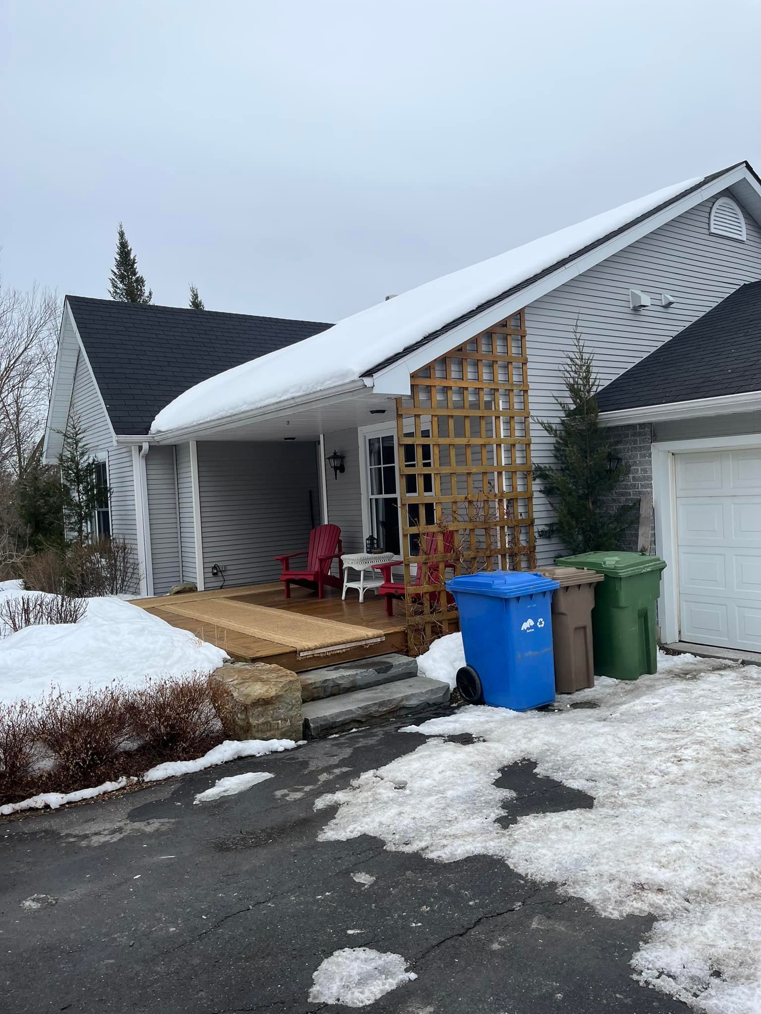 Extérieur d'une maison enneigée avec porche, terrasse et poubelles.