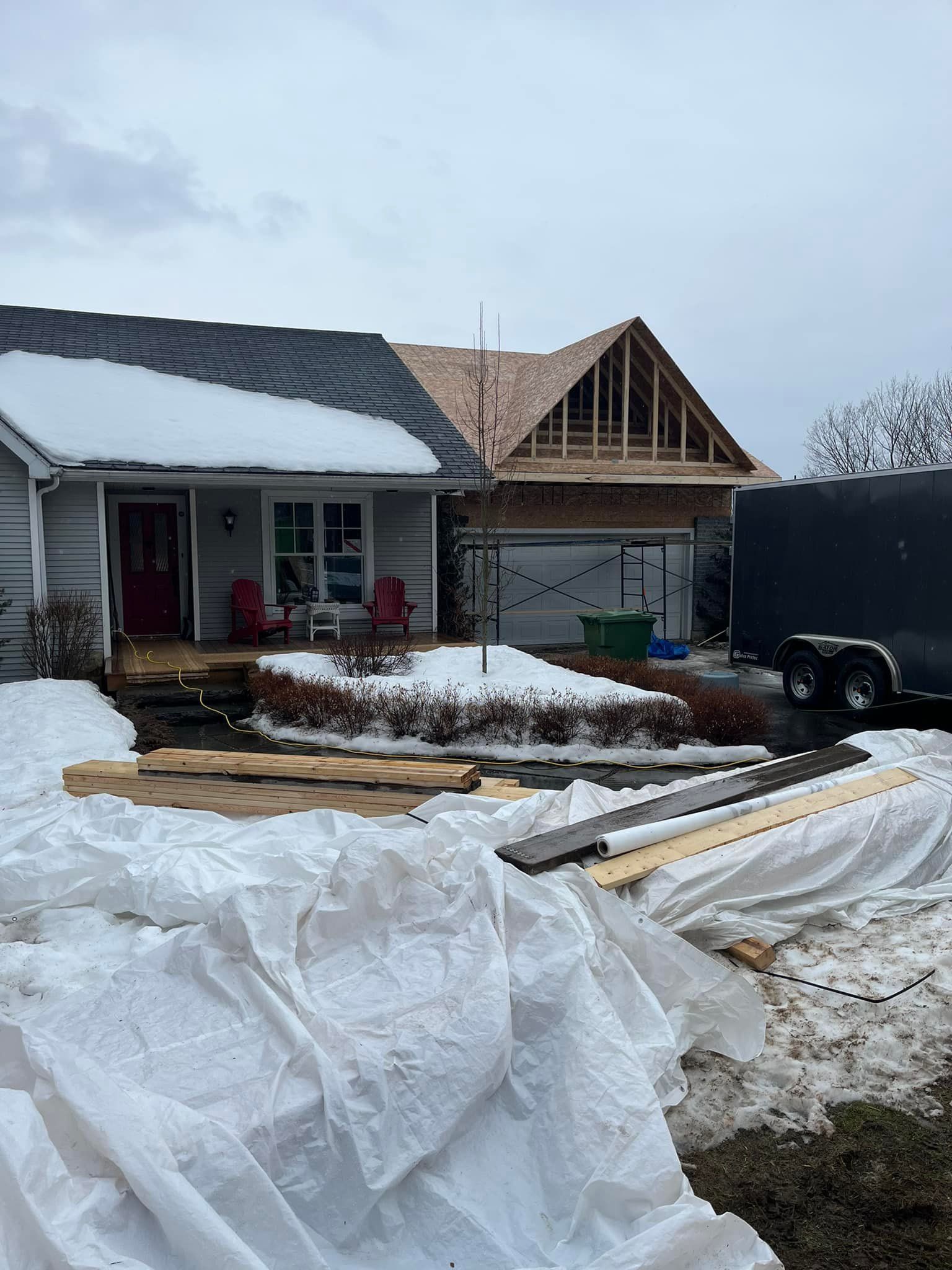 Maison en construction avec toit enneigé ; extension de garage inachevée. Bois de construction et bâche au premier plan.