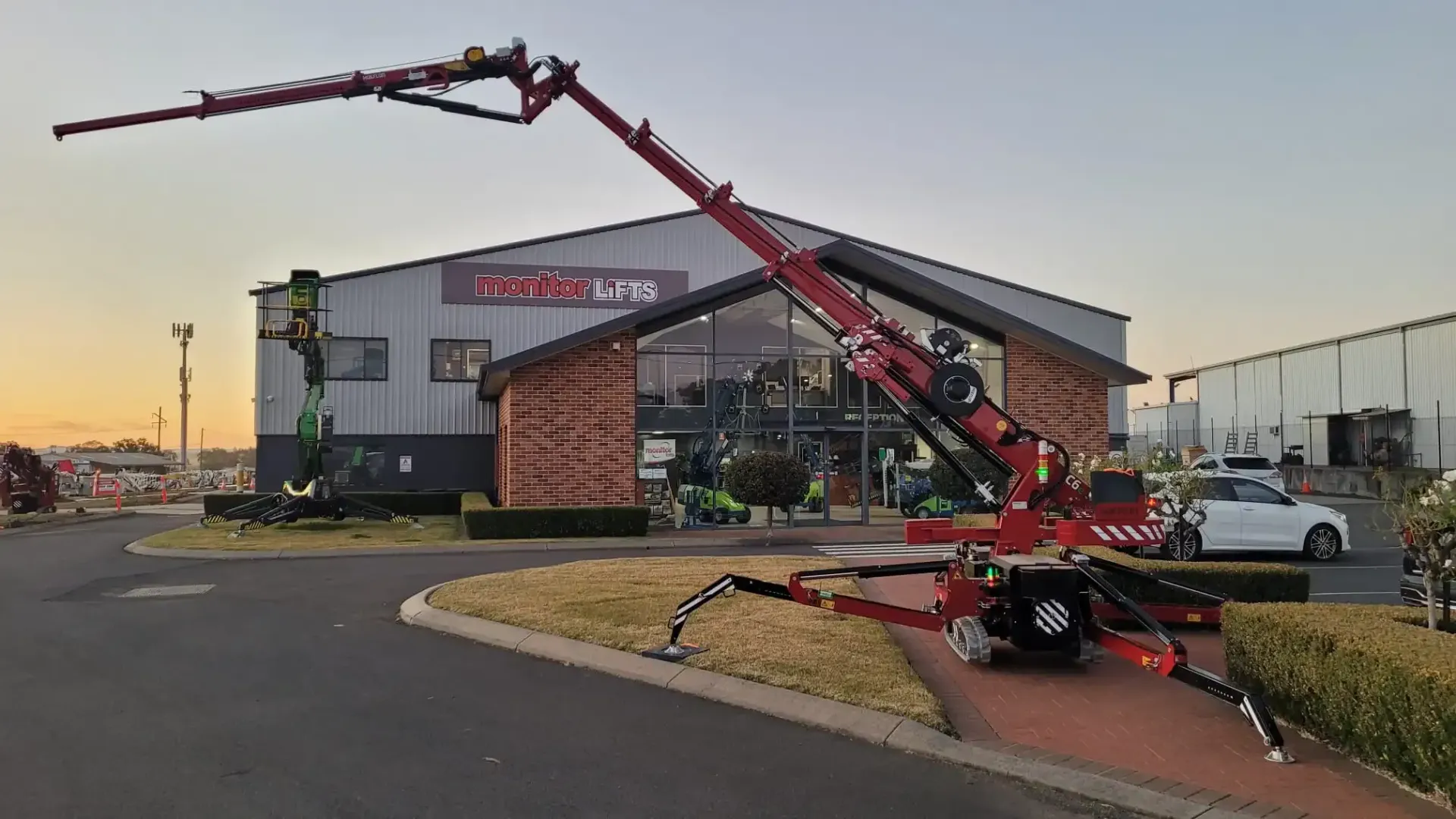 Red spider crane extended in front of a building with a glass front; parked on asphalt.