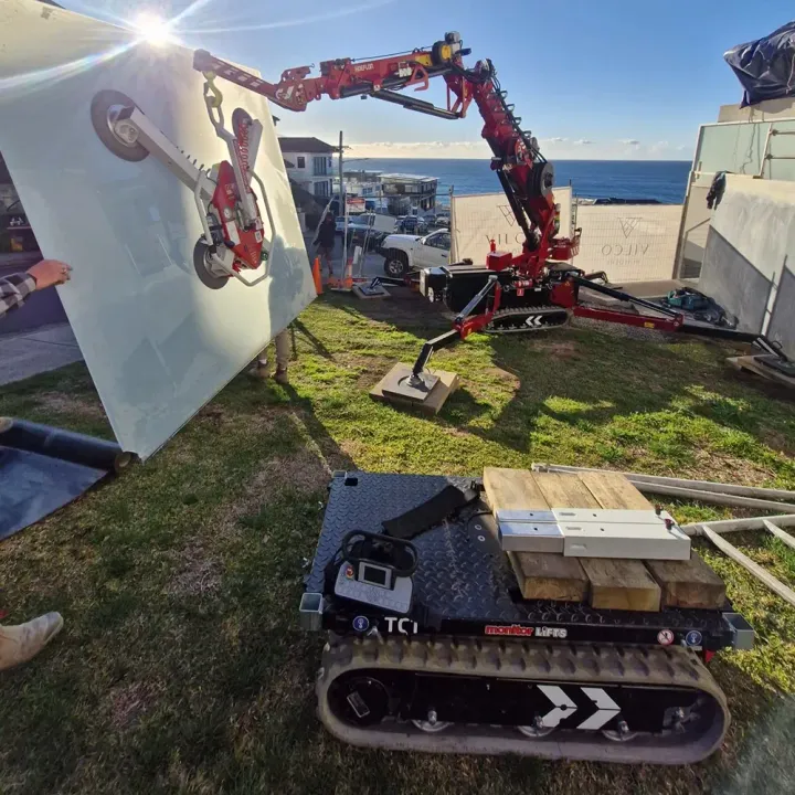 Red crane lifts large panel with tracked lift on grass near building and ocean.