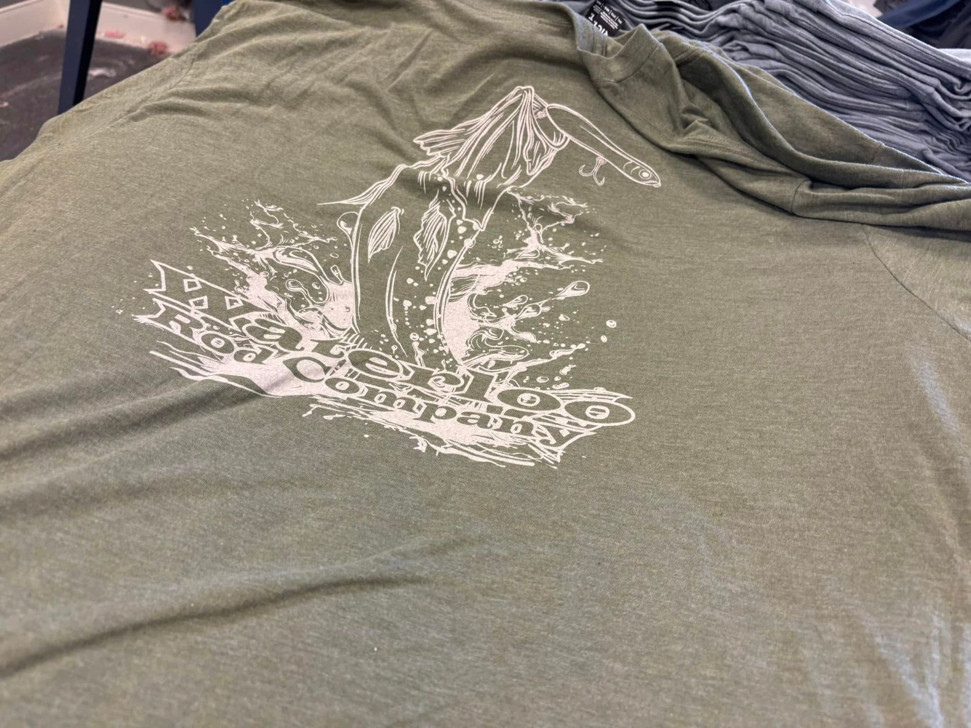 Olive green t-shirt with white graphic of a jumping fish, mountain, and 