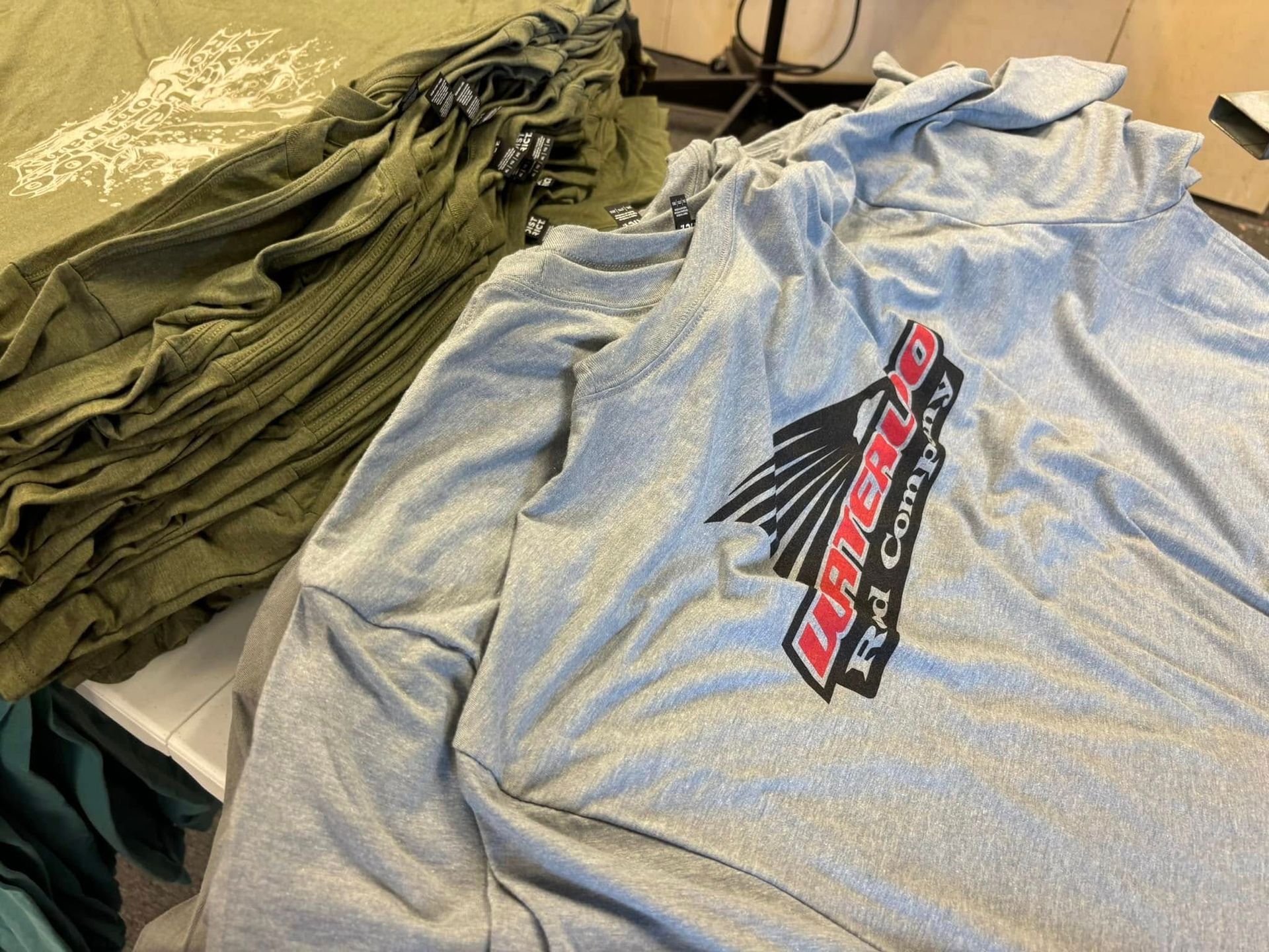 Stacks of olive green and gray t-shirts with logos, on a table.