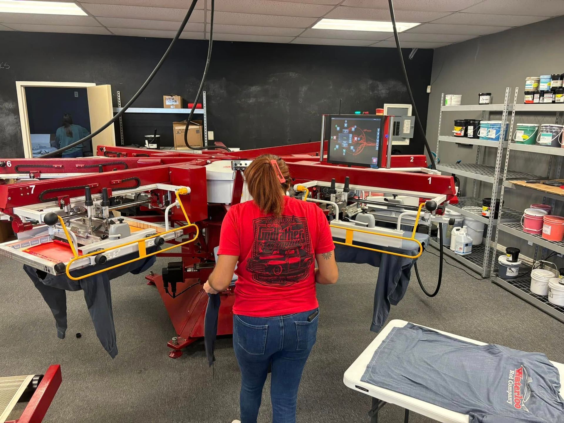 Woman in red shirt operates a screen printing machine; gray shirt on table, shelves with supplies in the background.