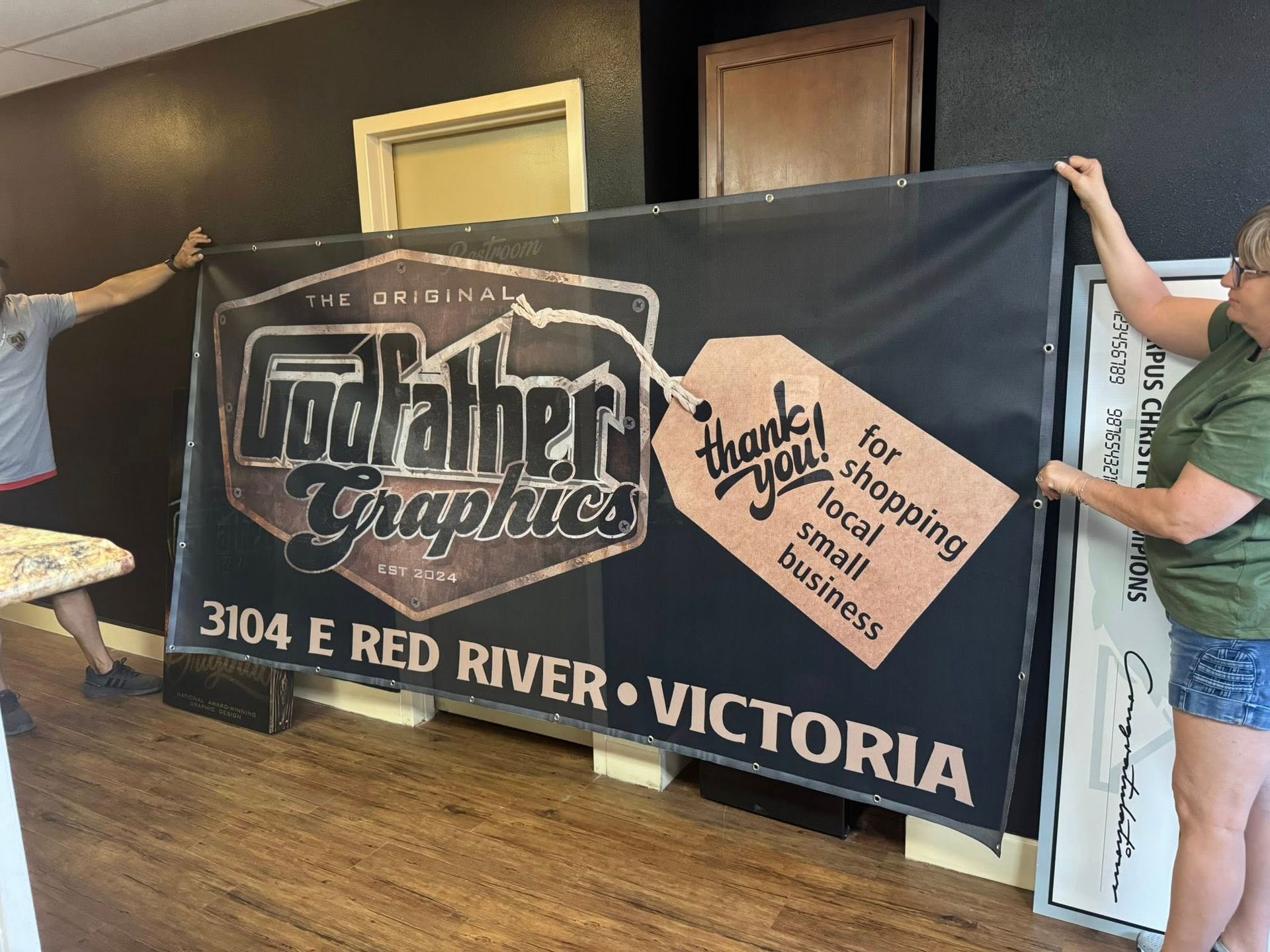 Two people holding a large banner for Godfather Graphics, thanking customers for supporting small businesses. Dark banner, shop interior.