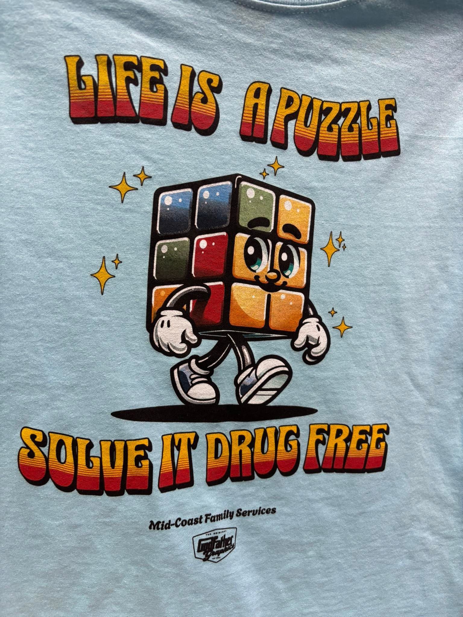 Light blue tee shirt with cartoon Rubik's cube person. Text says 