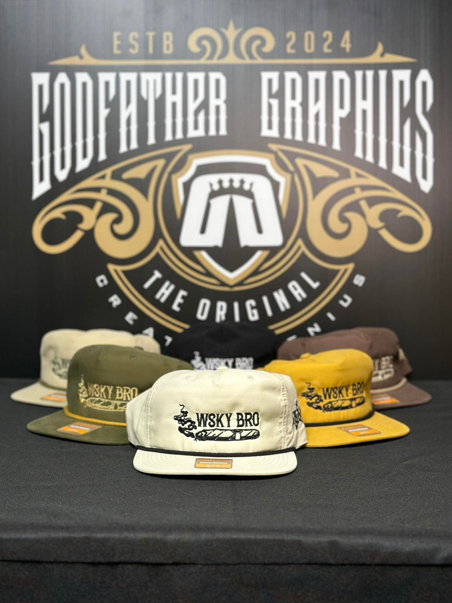 Godfather Graphics hats, various colors, displayed in front of logo on black background.