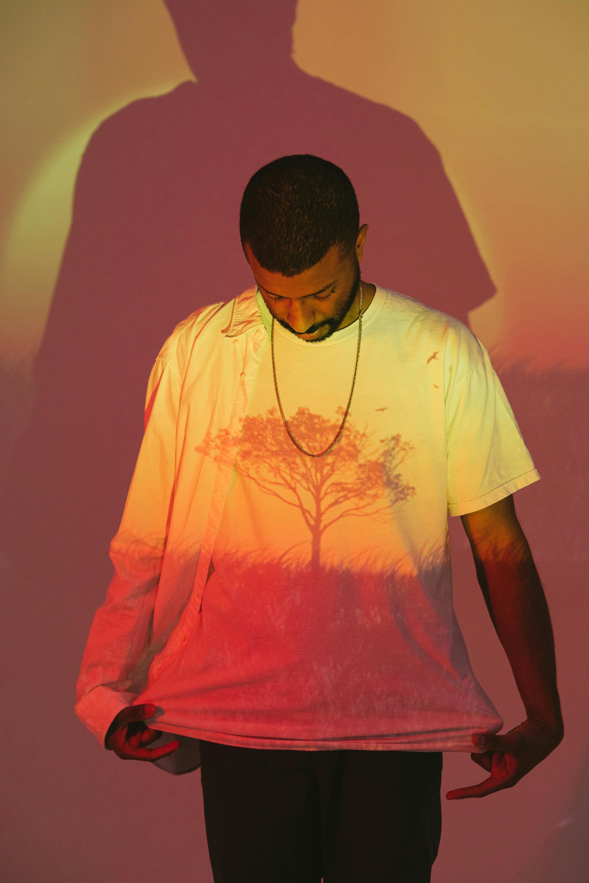 Man in white shirt with tree design, looking down. Lit with red/yellow, shadow behind.