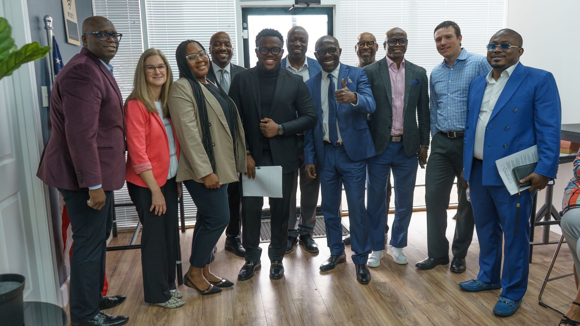 Group of people in business attire smiling in an office setting, some holding papers.