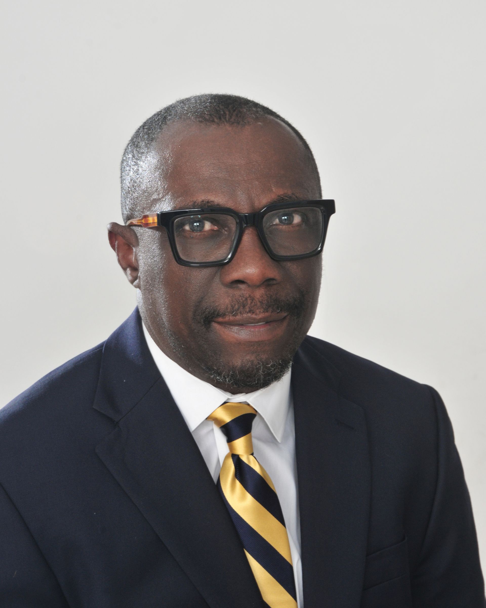 Woman in glasses watches a video call on a laptop and monitor, displaying a speaker with a government seal backdrop.