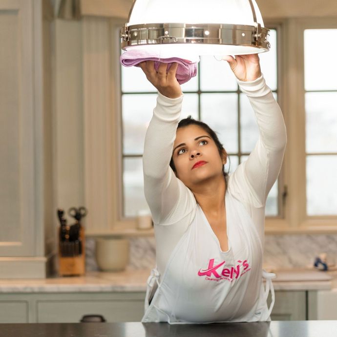 Woman cleaning a light fixture in a kitchen, wearing a white shirt and apron.