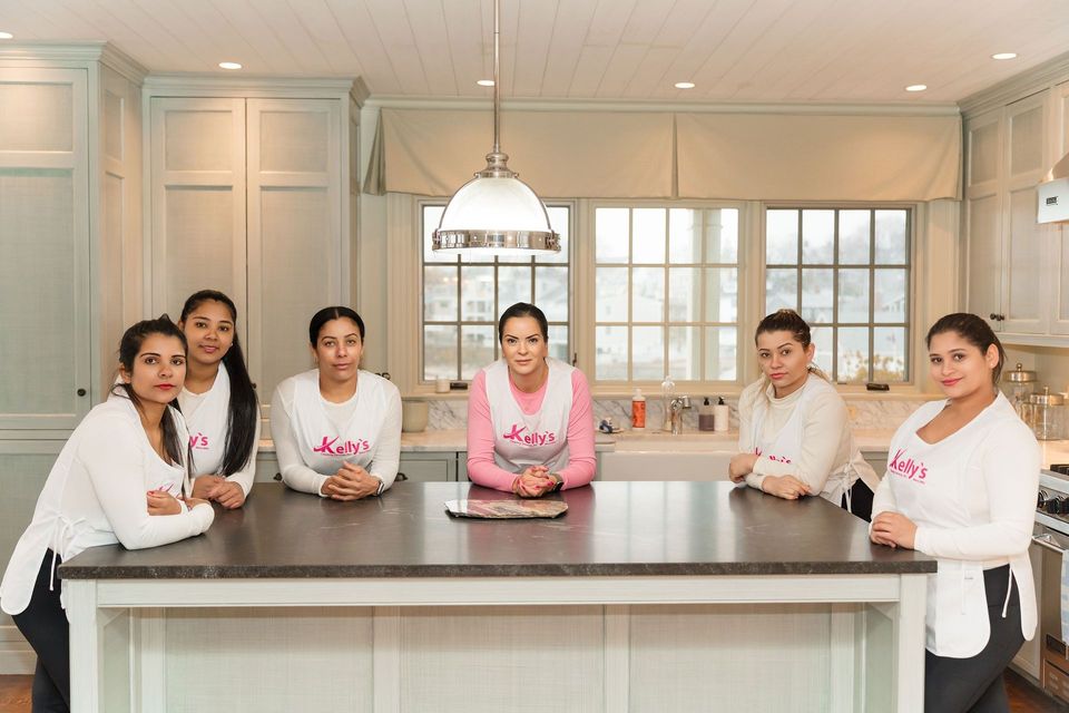 Seven women in a kitchen pose behind a dark countertop; some wear pink aprons.