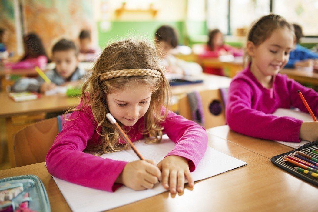 A girl in a pink shirt is writing in a notebook in a classroom.