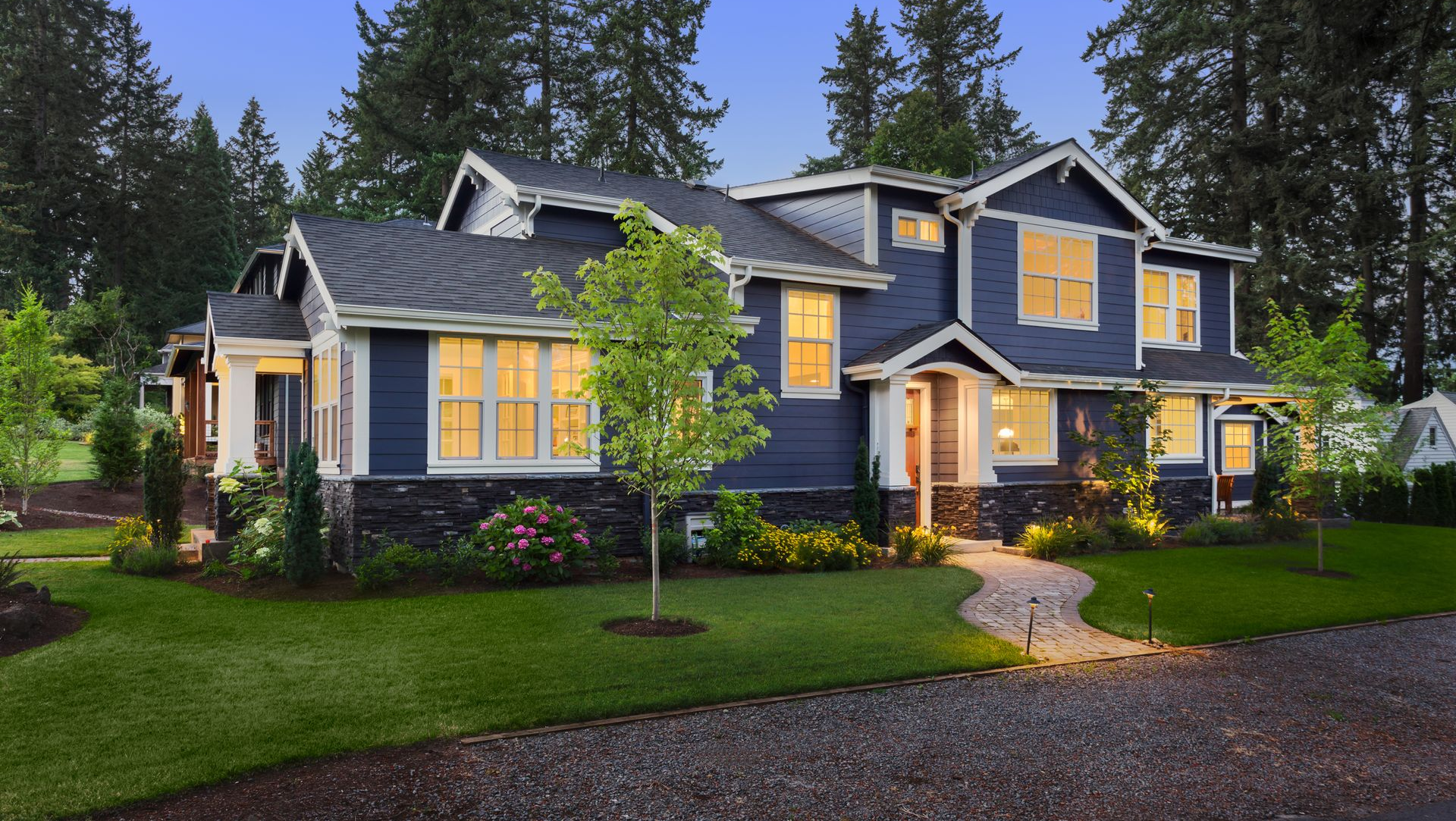 Blue two-story house with white trim, lit windows, and a stone walkway. Surrounded by trees and green lawn.