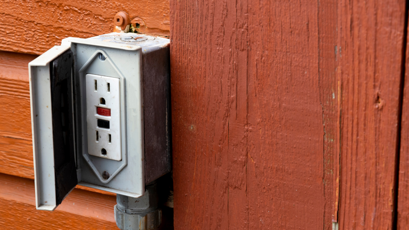 Gray weatherproof electrical outlet box mounted on red wooden siding.