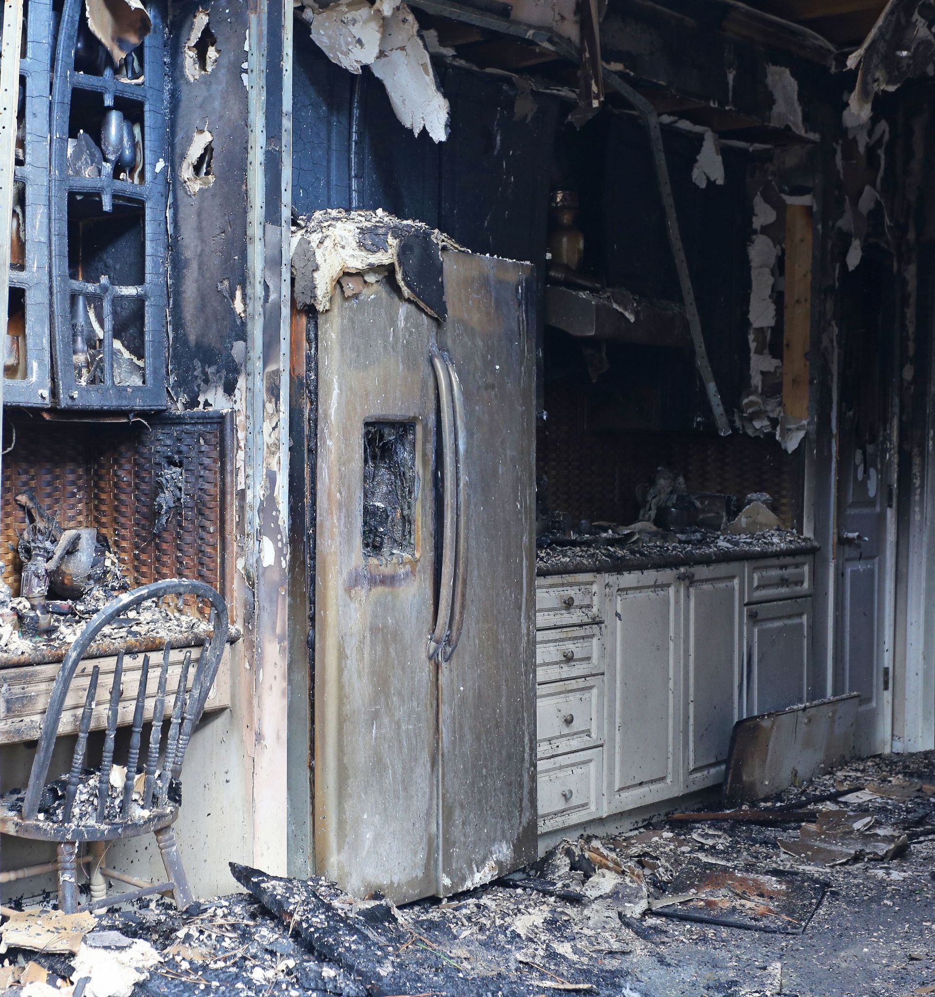 Kitchen destroyed by fire; refrigerator, cabinets, and walls are charred black. Debris covers the floor.