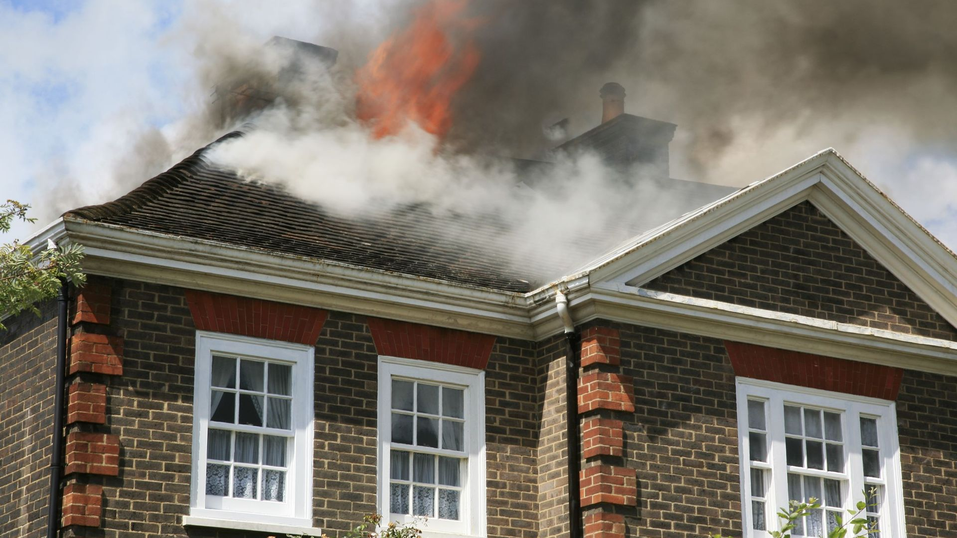 House engulfed in flames, thick black smoke billowing from the roof, orange fire visible.