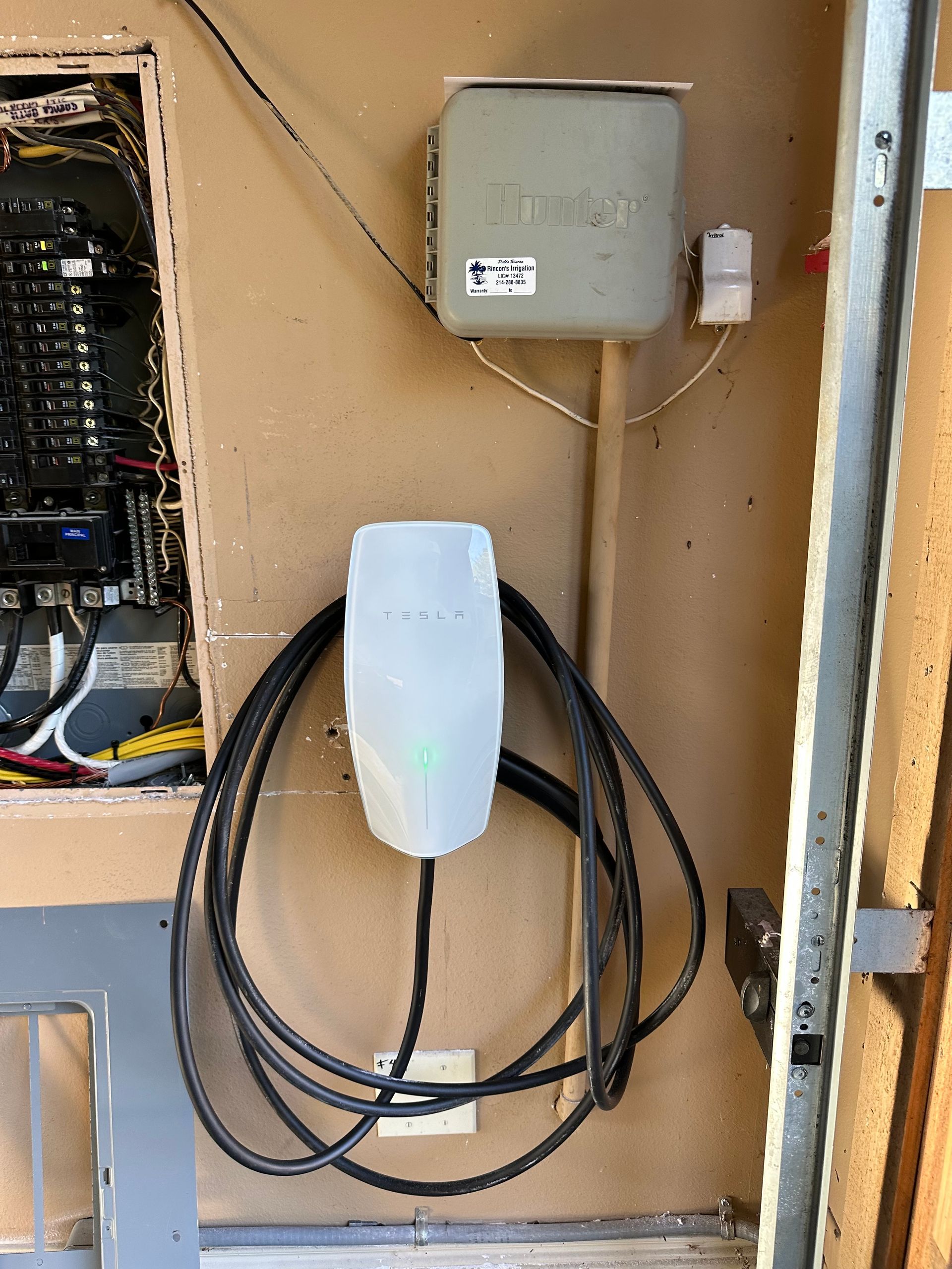 Electric vehicle charger mounted on a wall near a breaker panel. Black charging cable loops down.