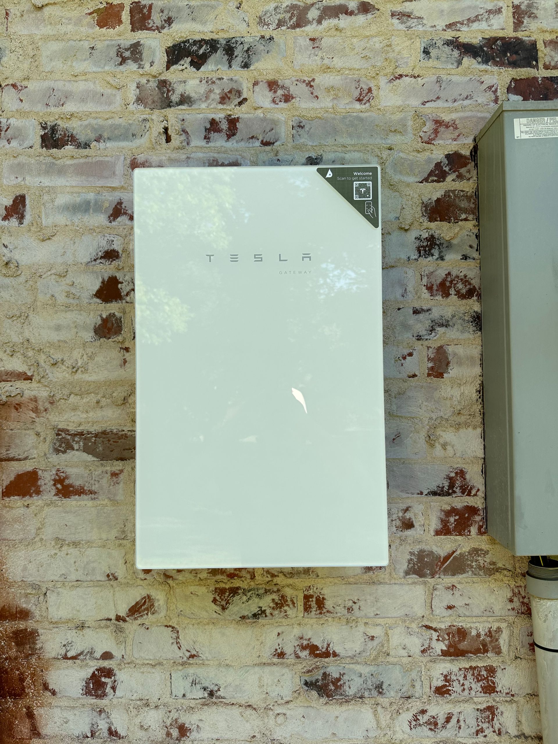 White Tesla Powerwall battery mounted on a weathered brick wall.