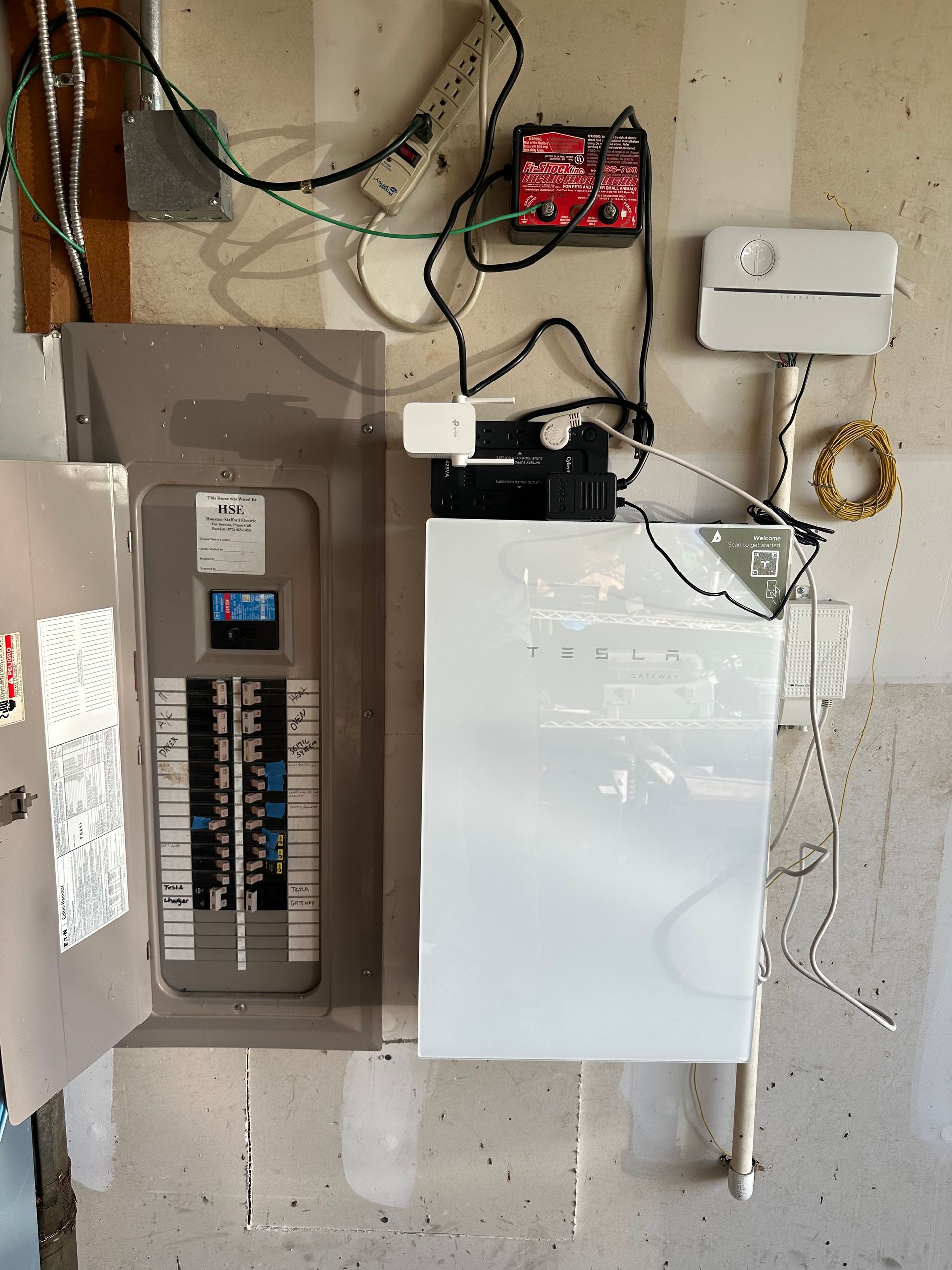 Electrical panel with circuit breakers, connected to white battery unit and other wiring on a wall.
