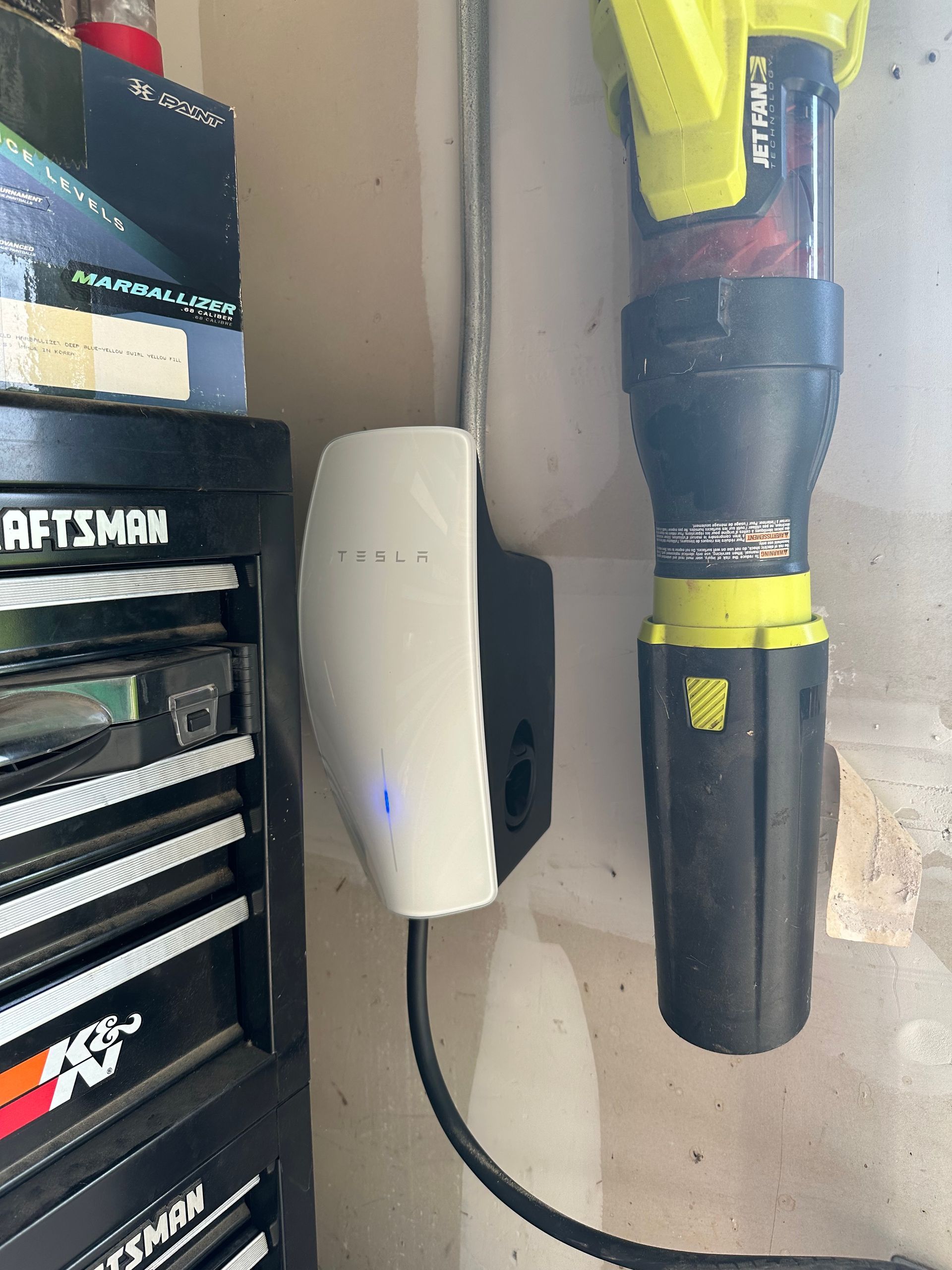 Tesla electric vehicle charger mounted on a garage wall next to a tool chest and a leaf blower.