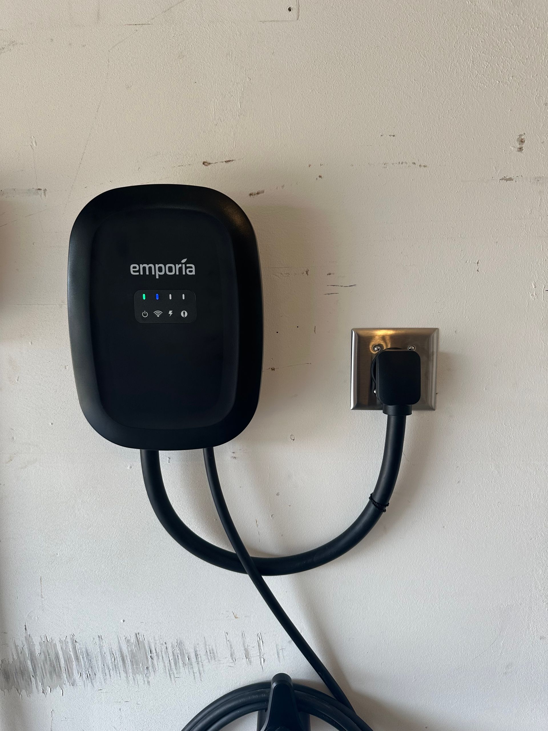 Black Emporia EV charger plugged into a wall outlet on a white wall.