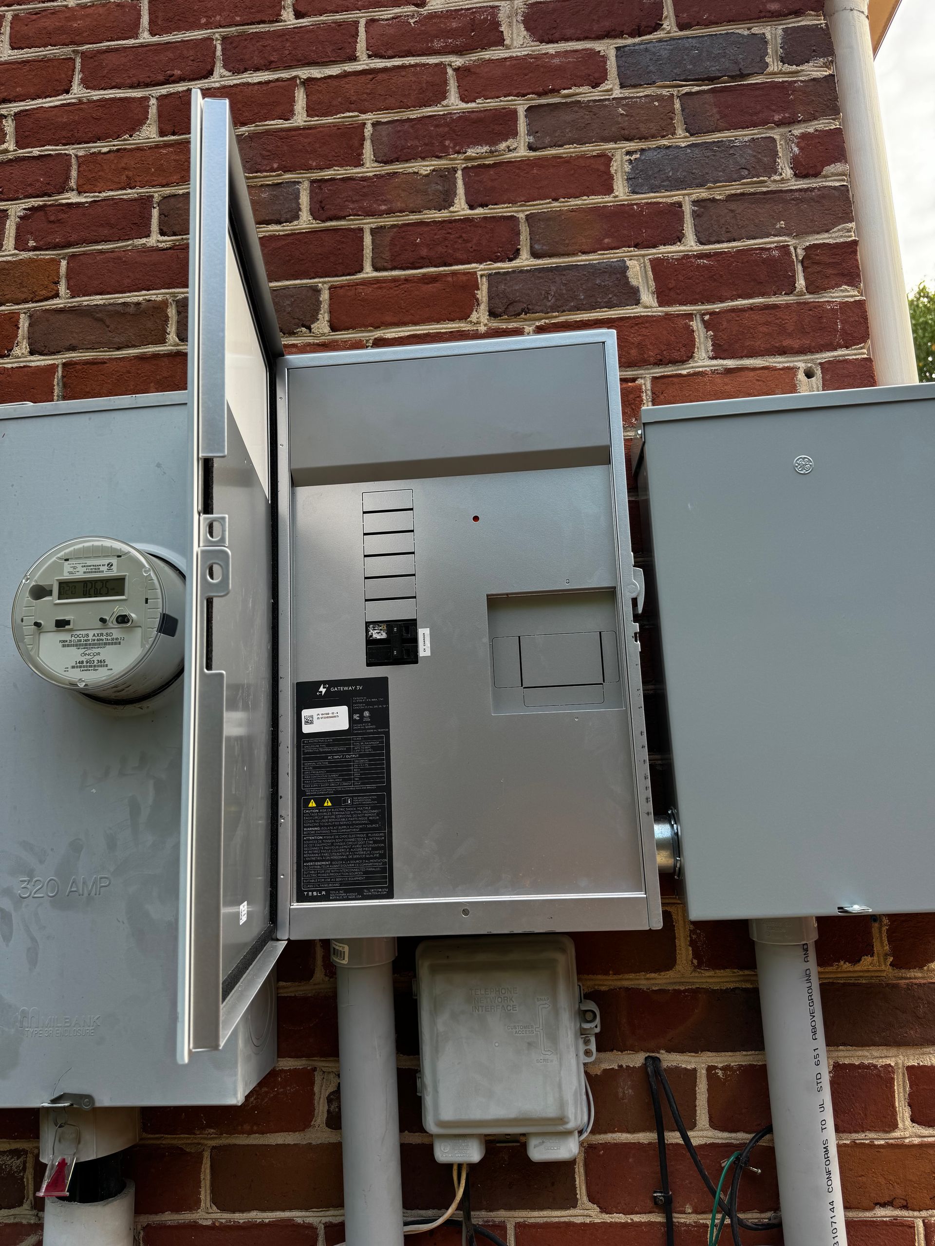Electrical meter and boxes mounted on a brick wall; one box door is open.