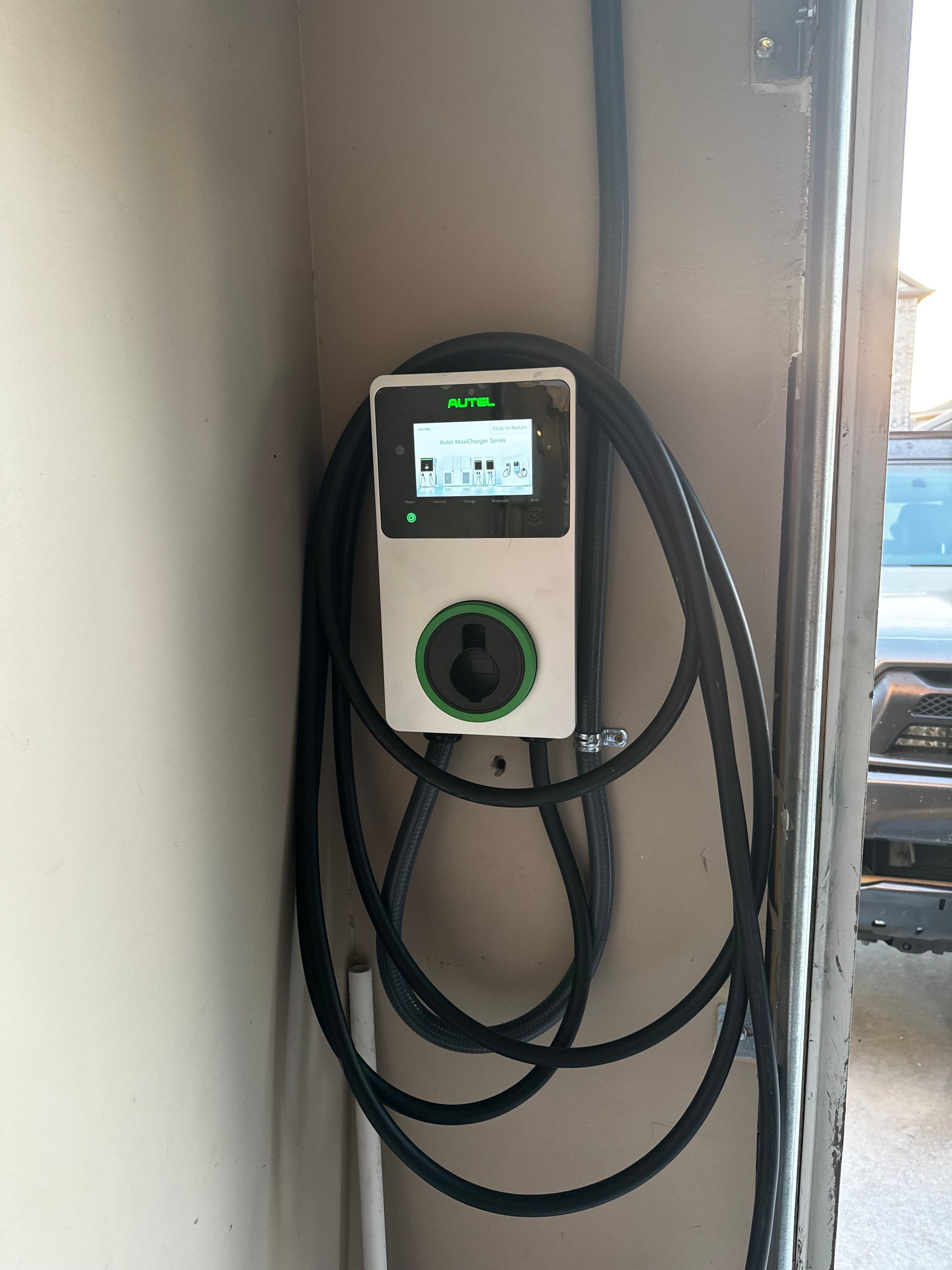 Electric vehicle charger mounted on a wall with a looped cable, screen illuminated.