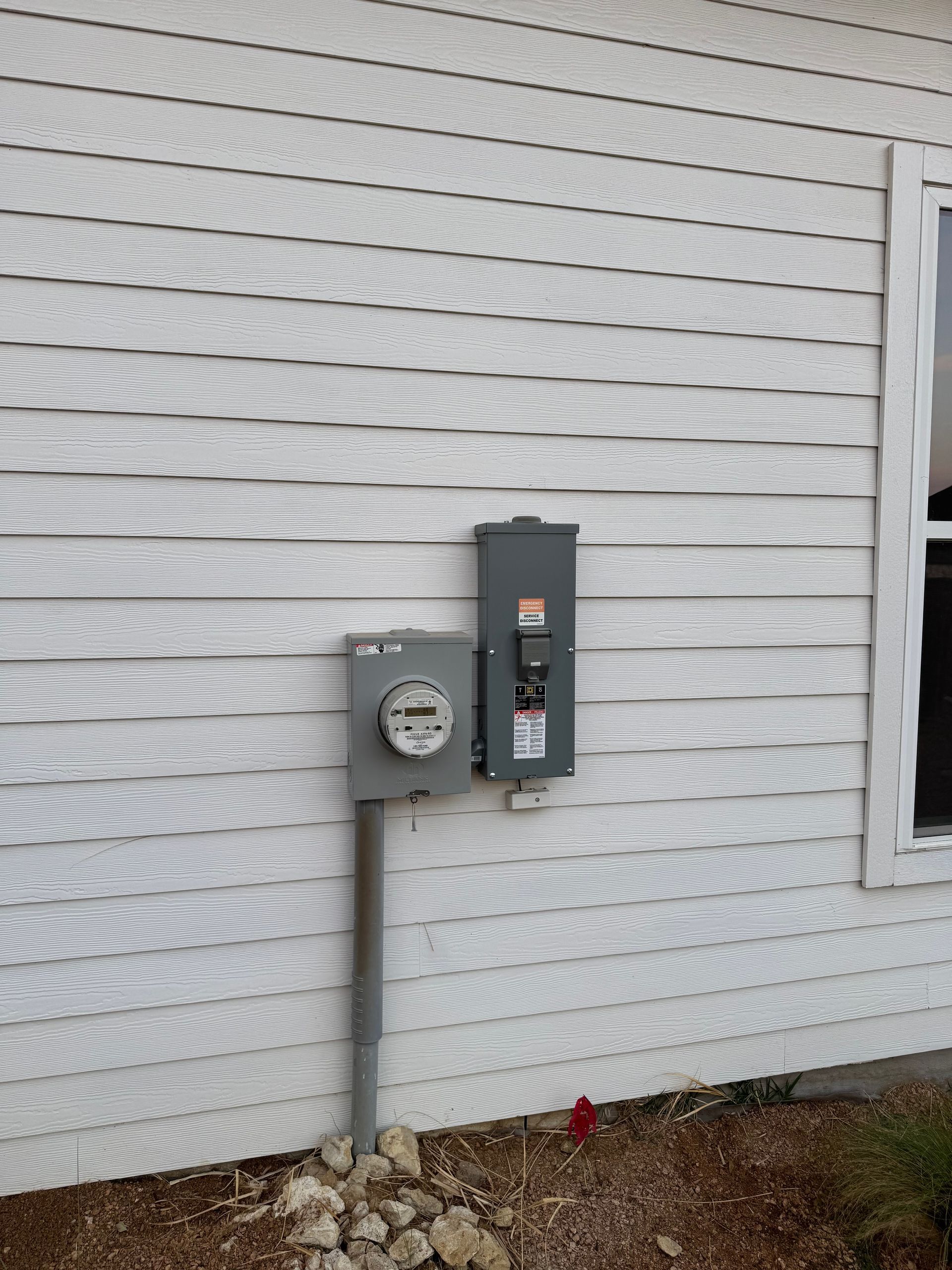 Electrical meter and circuit breaker box on the side of a white-sided building.