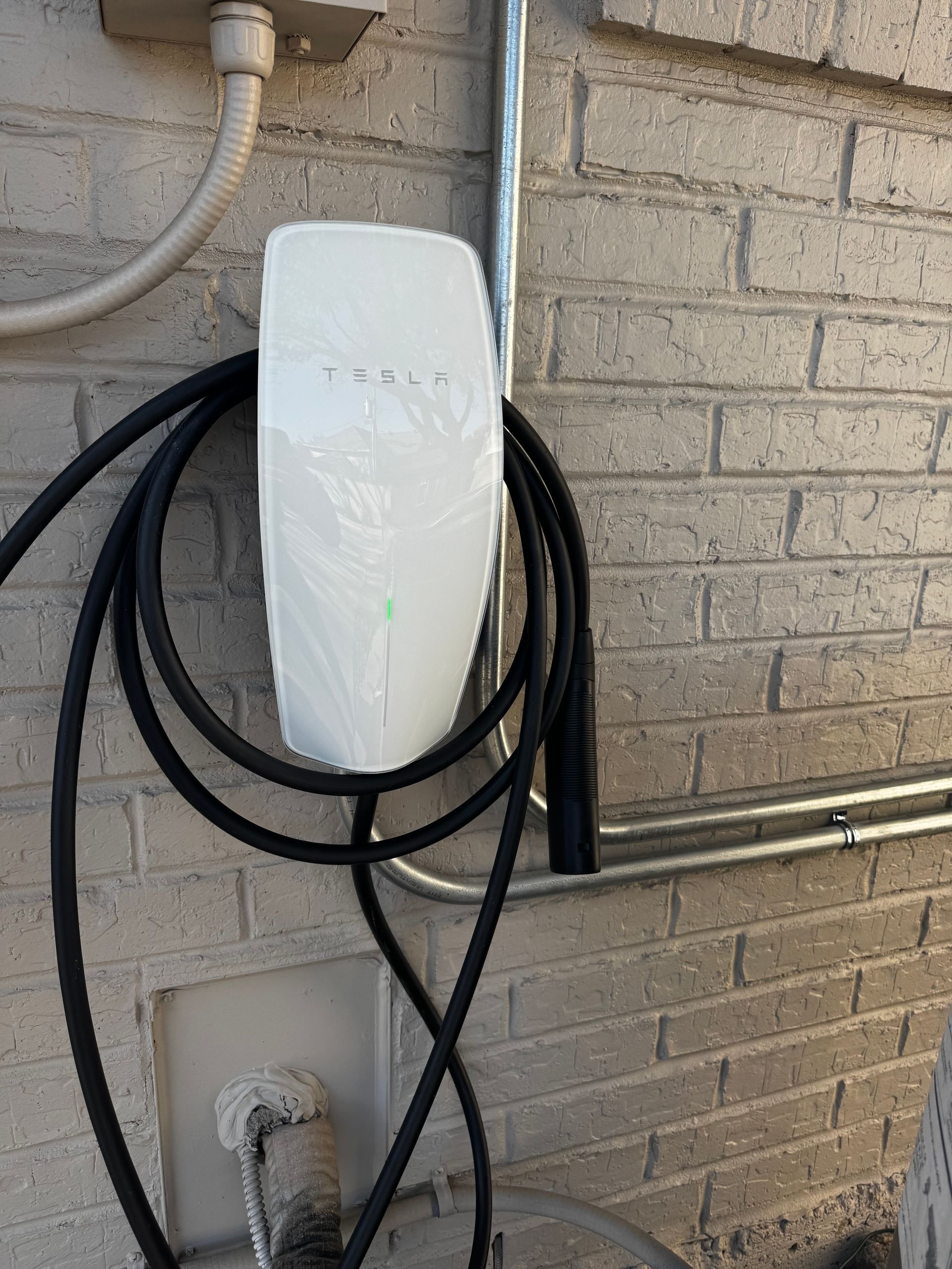 White Tesla wall charger mounted on a brick wall, with charging cable coiled below.