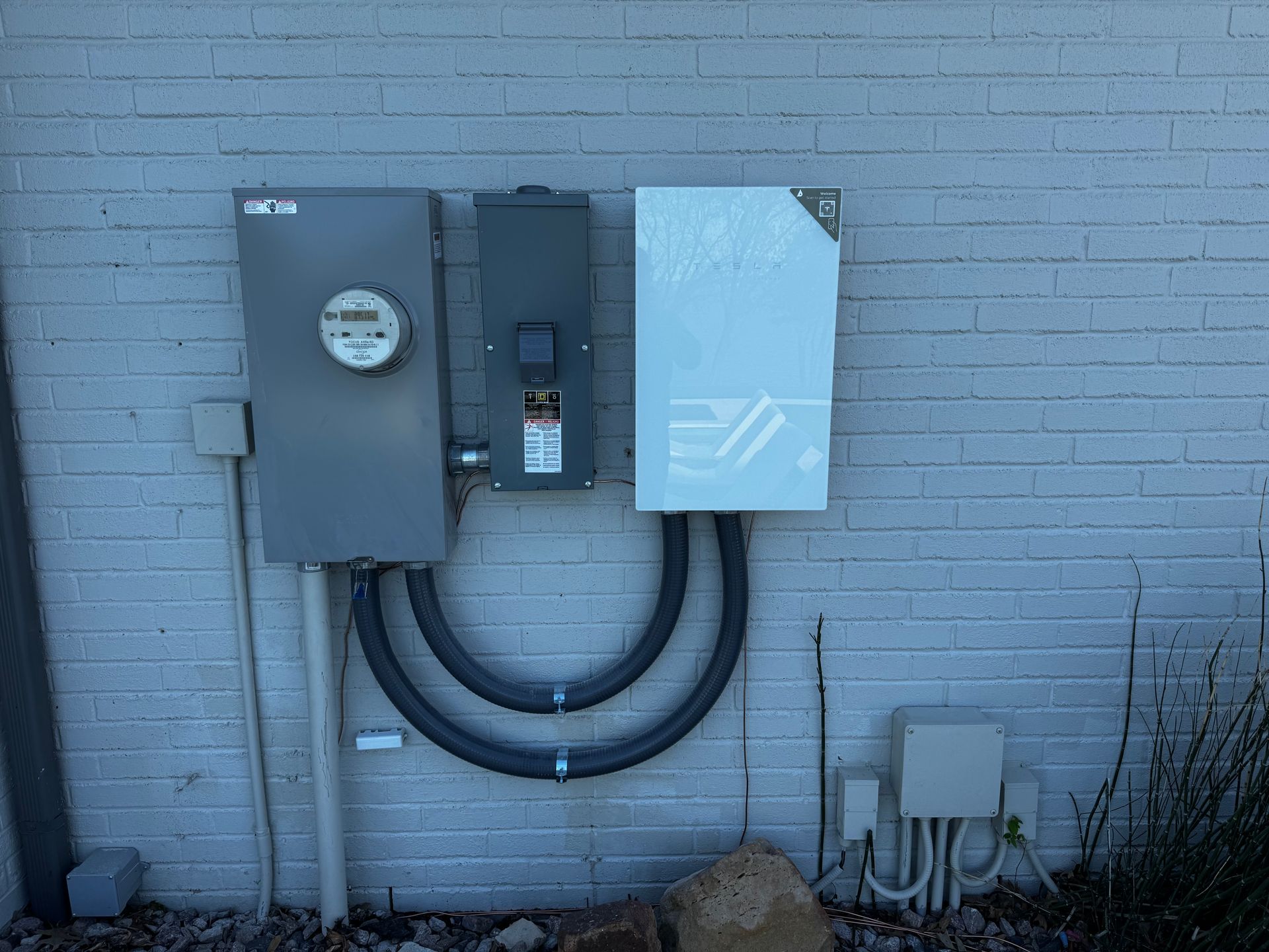 Solar panel equipment mounted on a brick wall: electrical meter, disconnect, and inverter with conduit.
