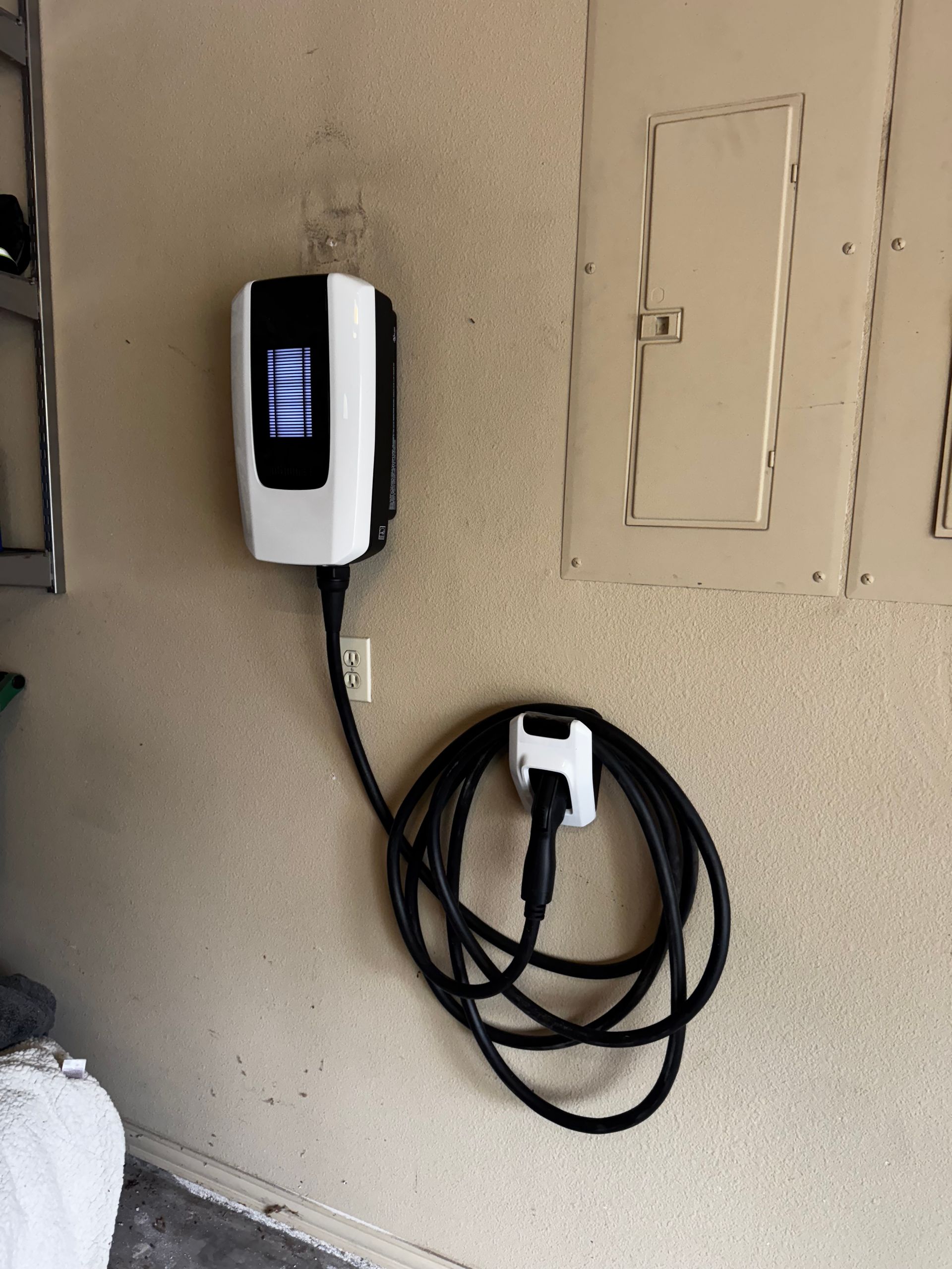 White EV charger mounted on a beige wall, cable coiled below. Next to a fuse box.