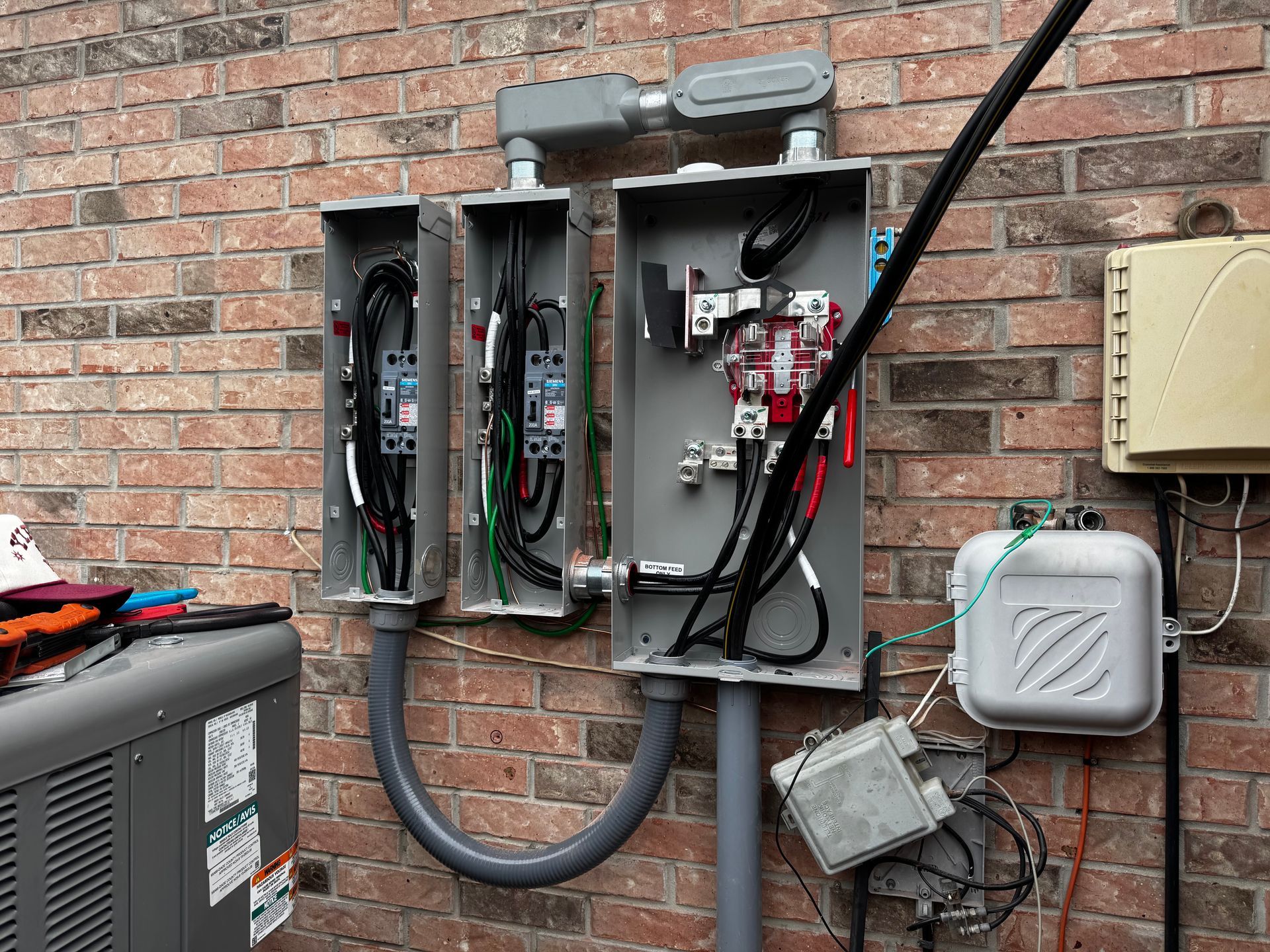 Electrical boxes mounted on a brick wall, with wires and conduits. A/C unit and utility box are visible.